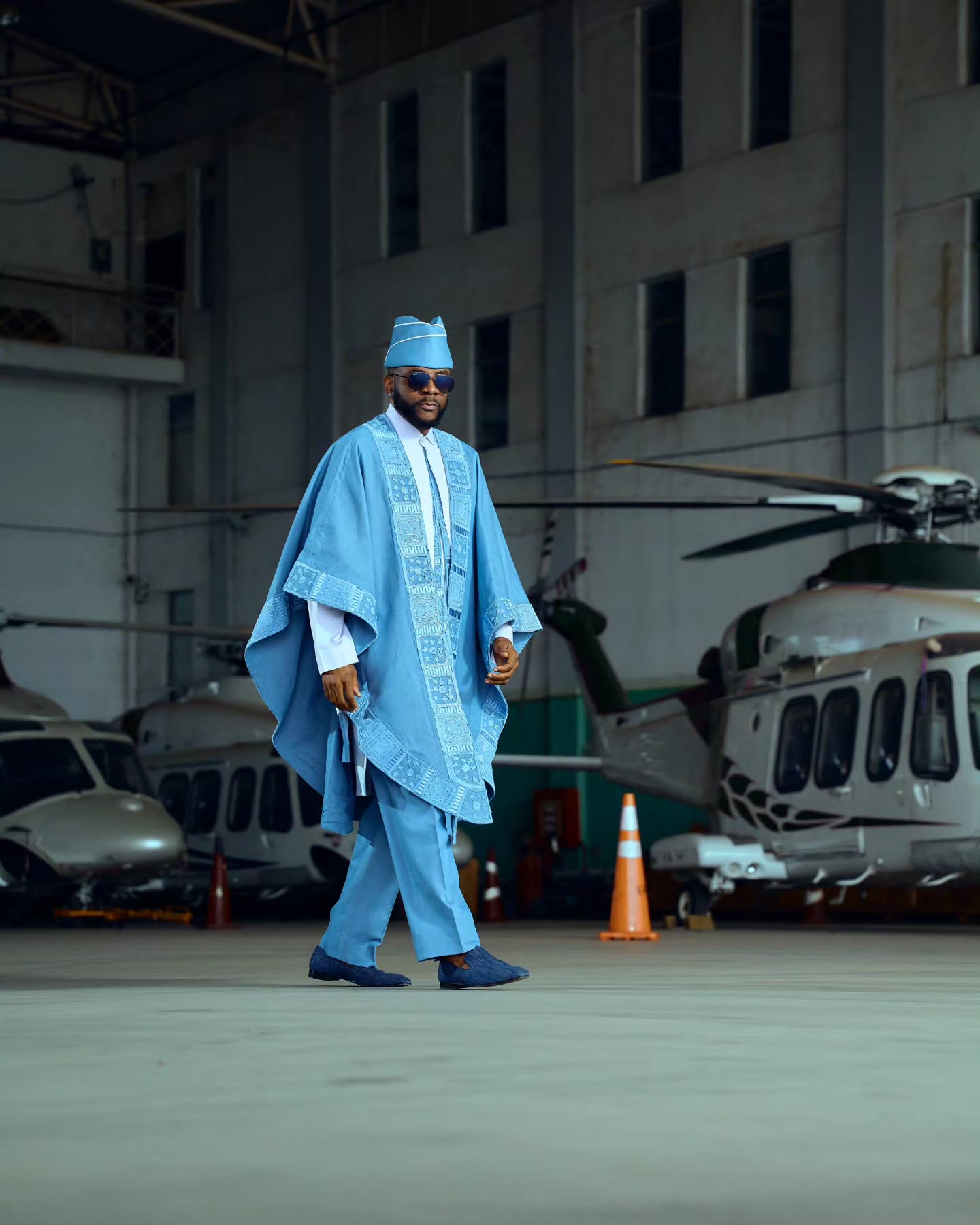 Ebuka Obi-Uchendu wearing a sky-blue denim Agbada by Deji and Kola with matching trousers, aviator sunglasses, and blue loafers inside an aircraft hangar.