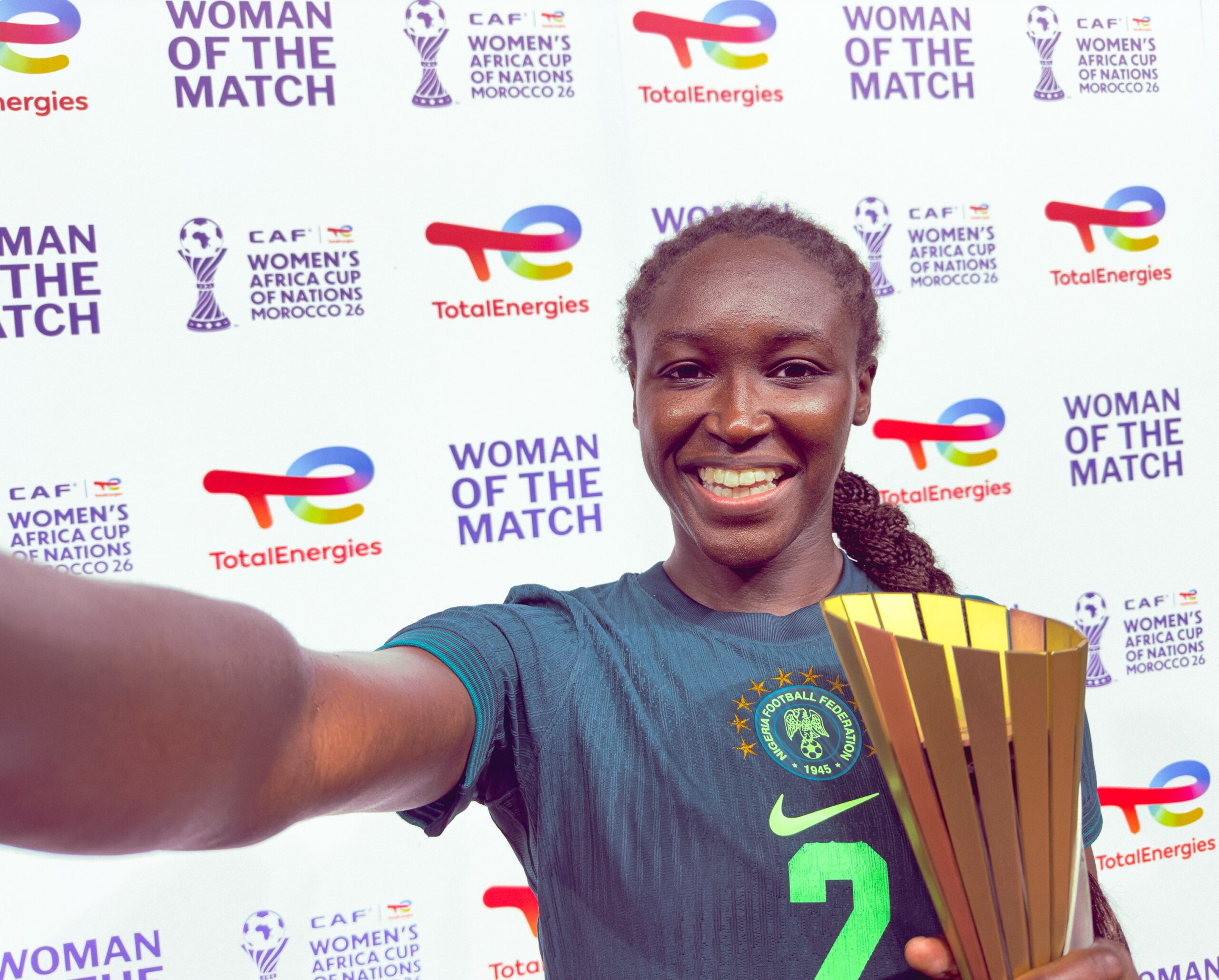 Rinsola Babajide taking a selfie holding the Woman of the Match trophy against the official WAFCON 2026 backdrop.