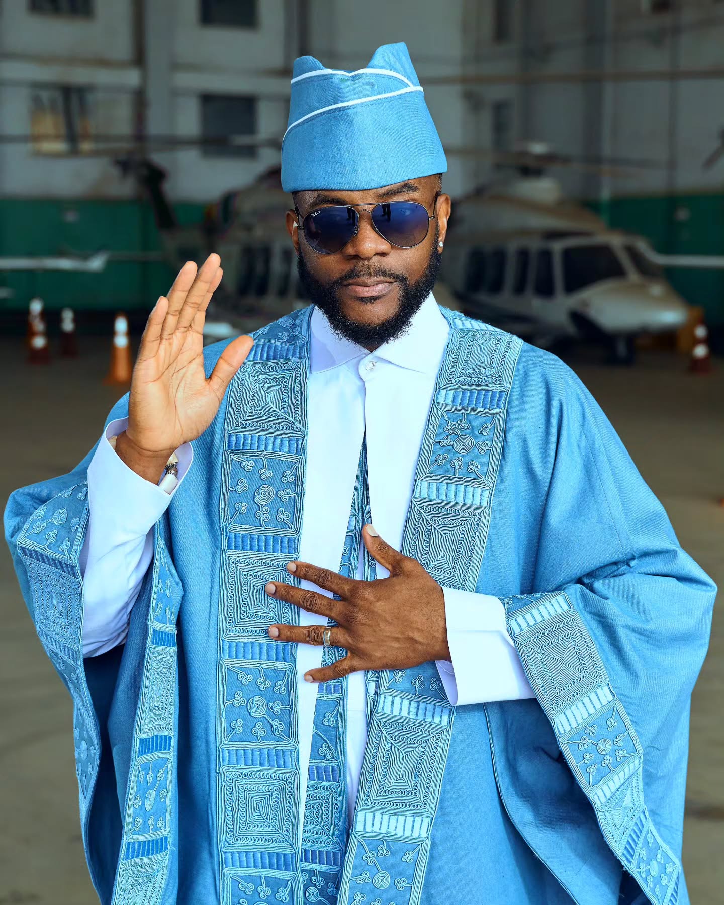 buka Obi-Uchendu gesturing in a sky-blue denim Agbada featuring intricate geometric embroidery down the front panels and a white collared Buba underneath.