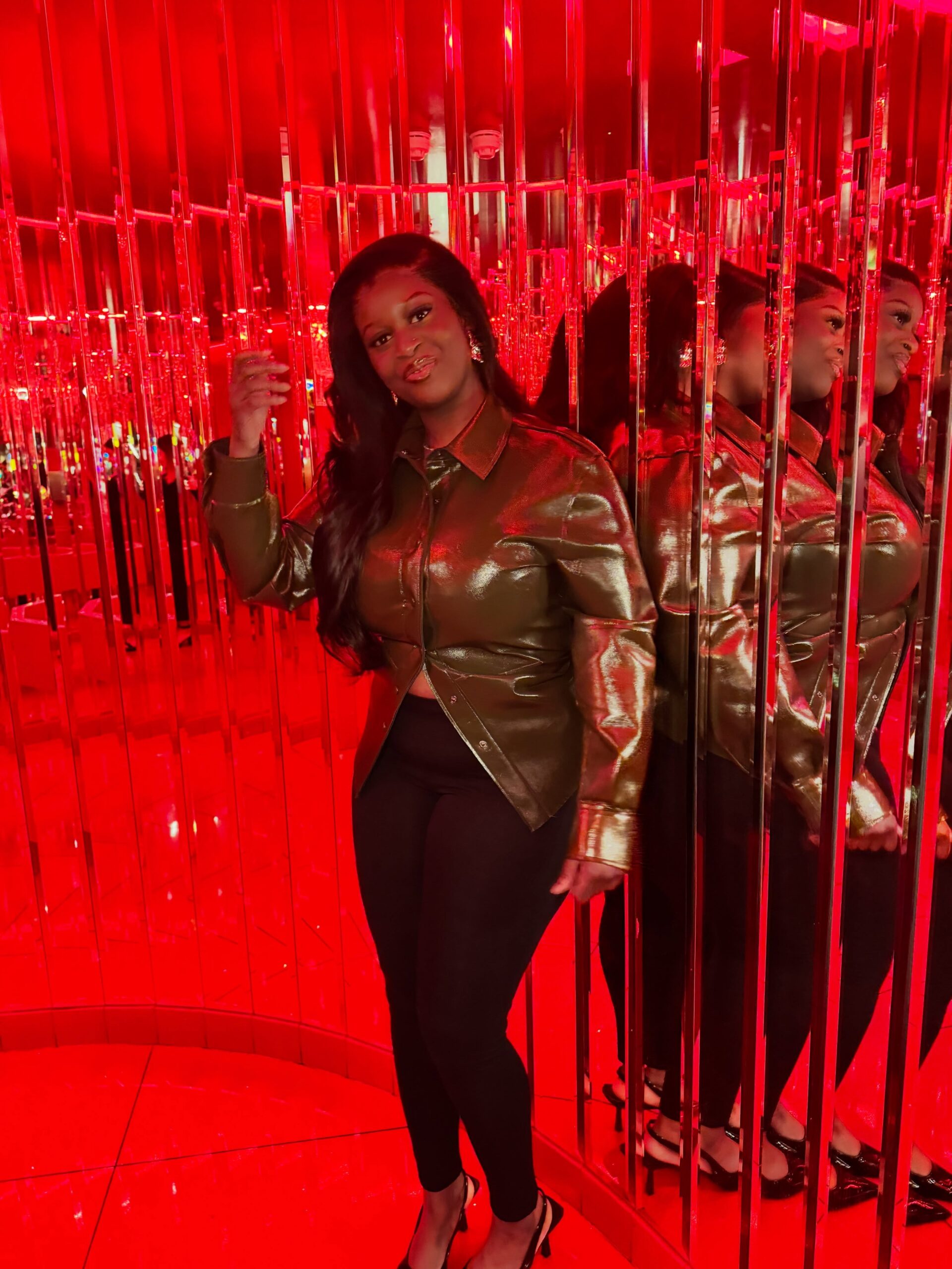 Temi Nkem wearing a metallic green shirt jacket, black trousers, and slingback heel shoes in a red mirrored room.