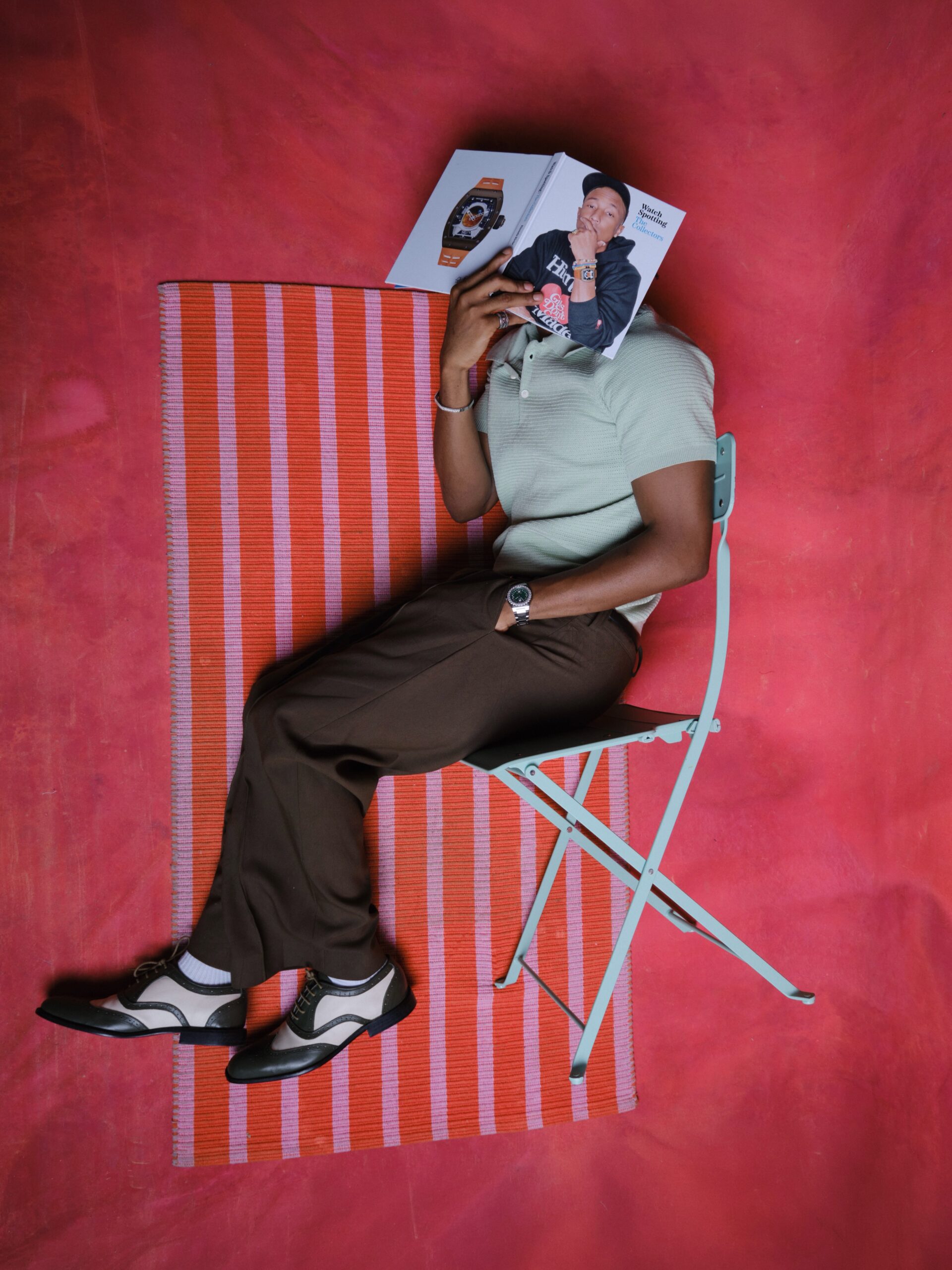 Mike Afolarin wearing a mint green short-sleeved knit polo, high-waisted olive brown wide-leg trousers, and two-tone spectator oxford shoes while holding a watch book.