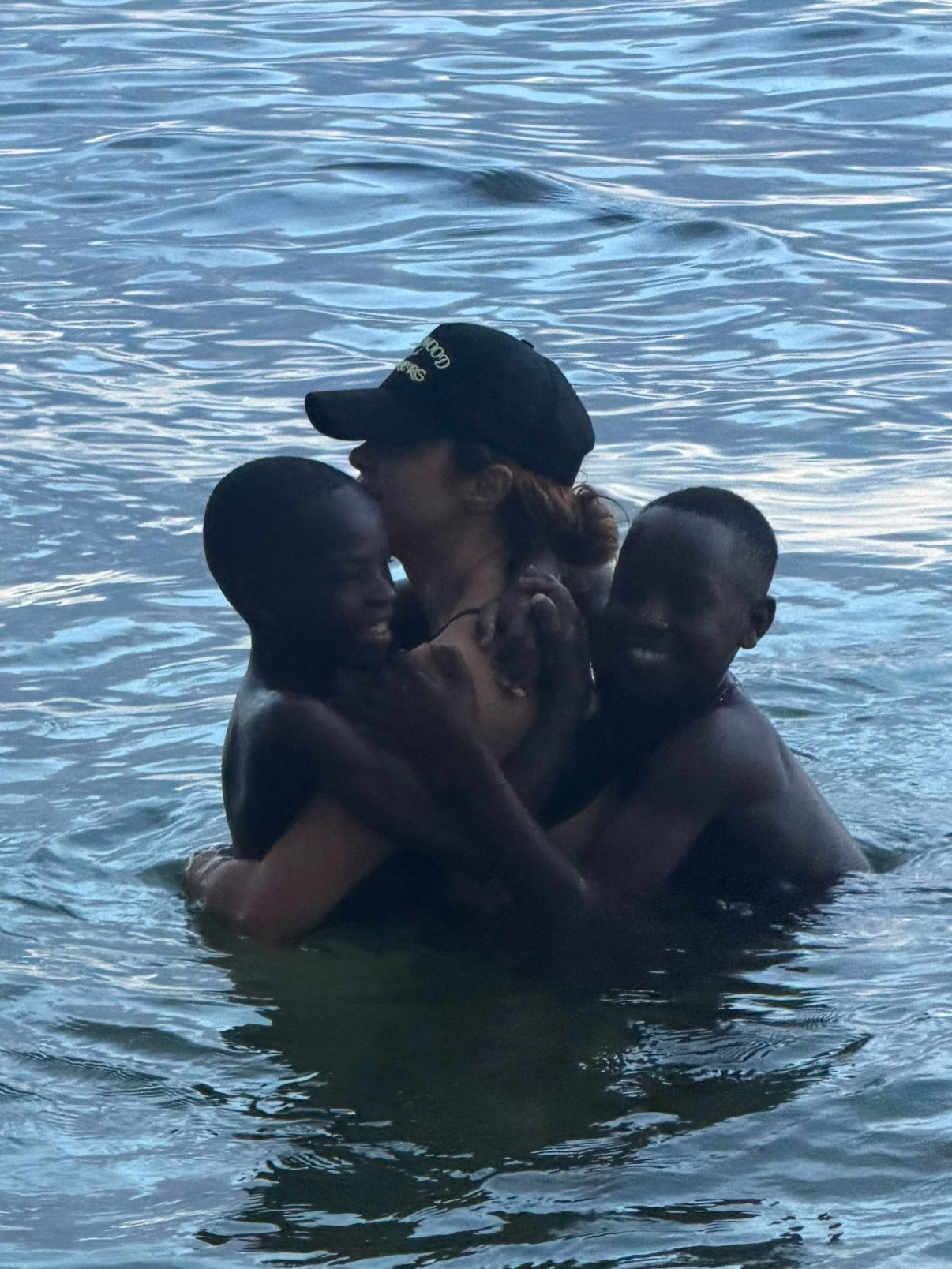 Shakira wearing a black cap while swimming in the ocean and hugging two members of the Triplets Ghetto Kids in the water.