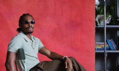 Mike Afolarin wearing a short-sleeved mint green knit polo, olive brown trousers, dark rectangular sunglasses, and two-tone oxford shoes.