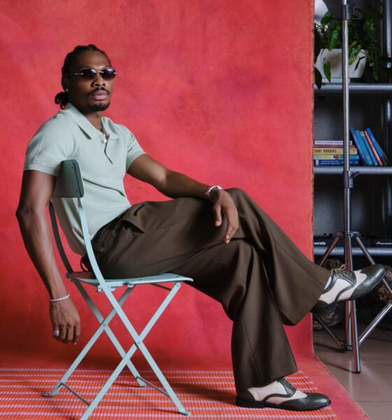 Mike Afolarin wearing a short-sleeved mint green knit polo, olive brown trousers, dark rectangular sunglasses, and two-tone oxford shoes.