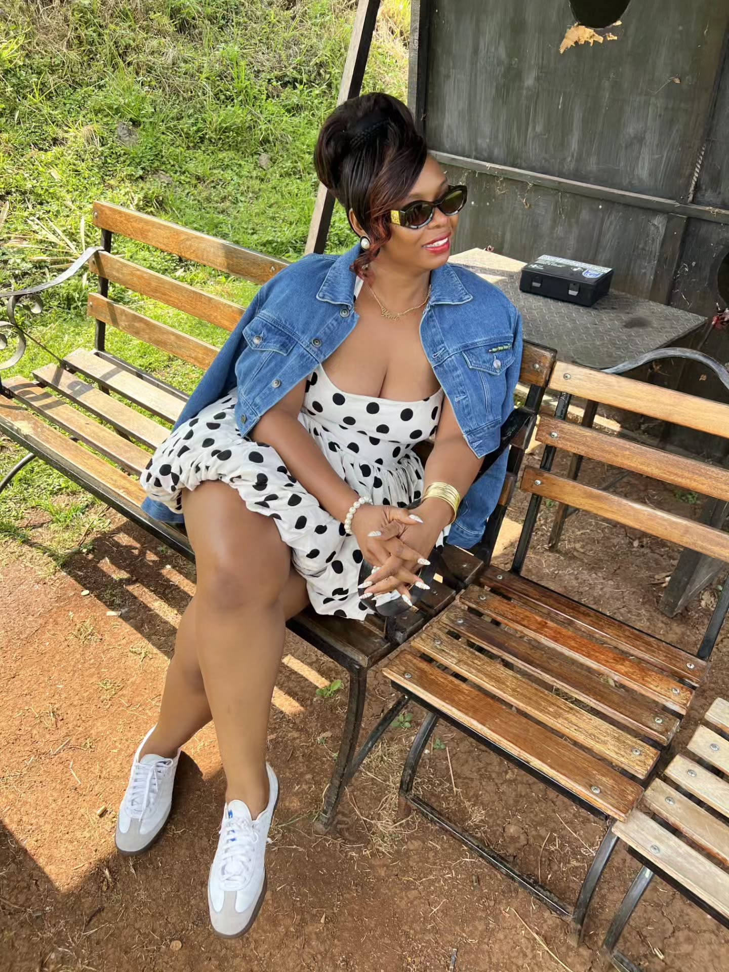 Yemi Alade sitting on a wooden bench wearing a white polka dot mini dress, denim jacket, and white low-top trainers.