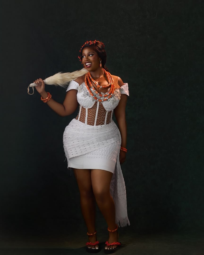 Temi Nkem wearing a white corset lace mini dress accessorised with coral beads and holding a traditional horsetail whisk.