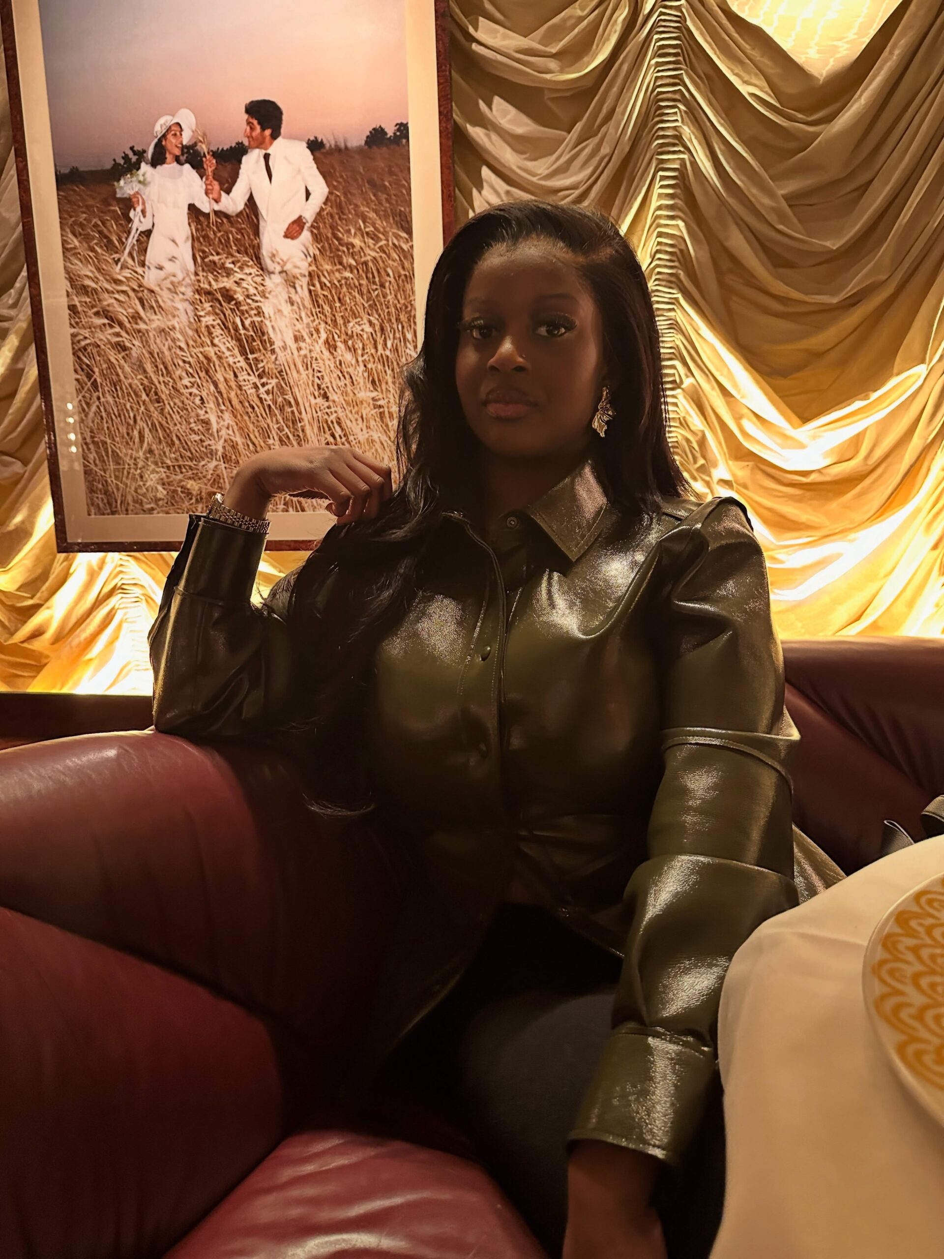 Temi Nkem wearing a metallic olive green button-down jacket seated on a red leather sofa.