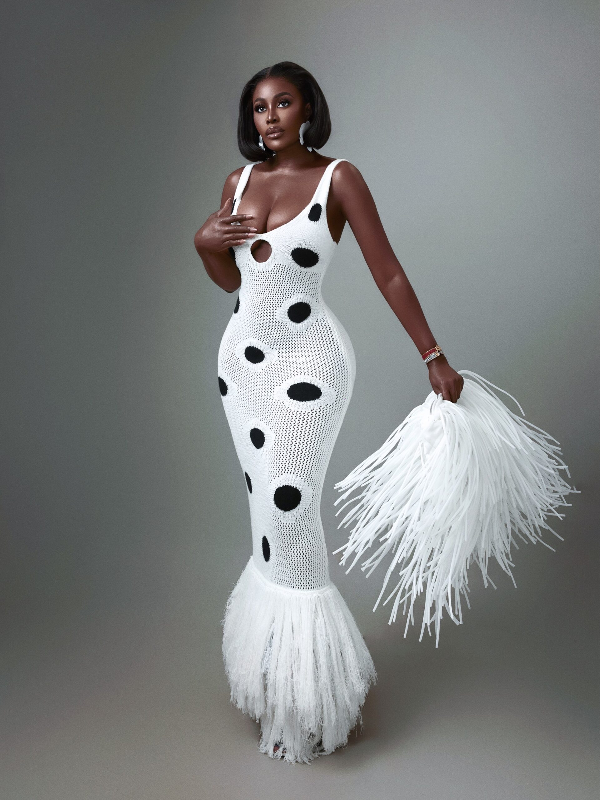 Ghanaian style icon Nana Akua Addo posing in a full-length white open-weave knit Hanifa dress with oversized black polka dots and a fringe hem, holding a white fringe clutch purse.