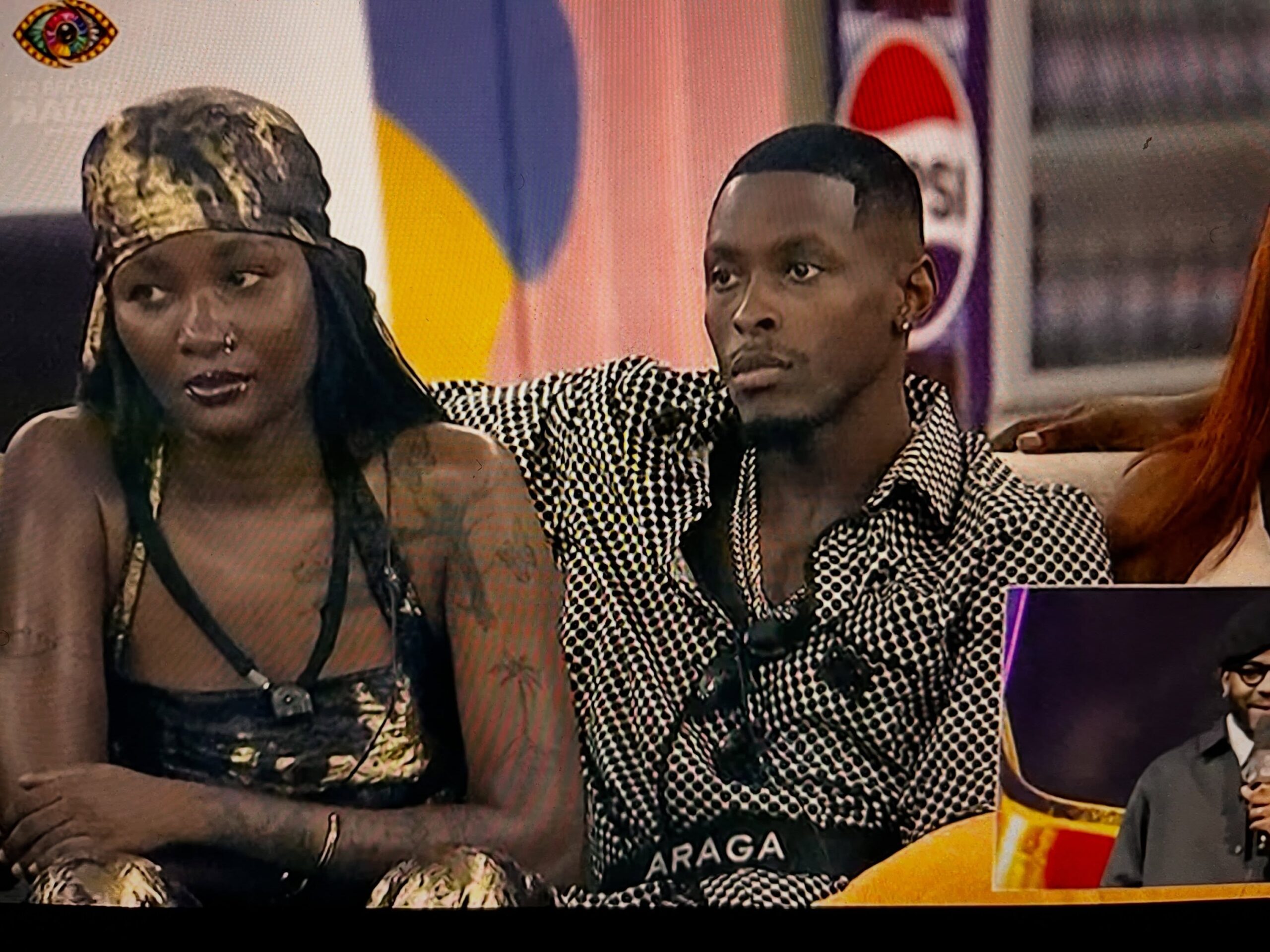 BBNaija housemate Araga in a patterned shirt sitting next to a female housemate wearing a patterned headscarf on the lounge sofa.