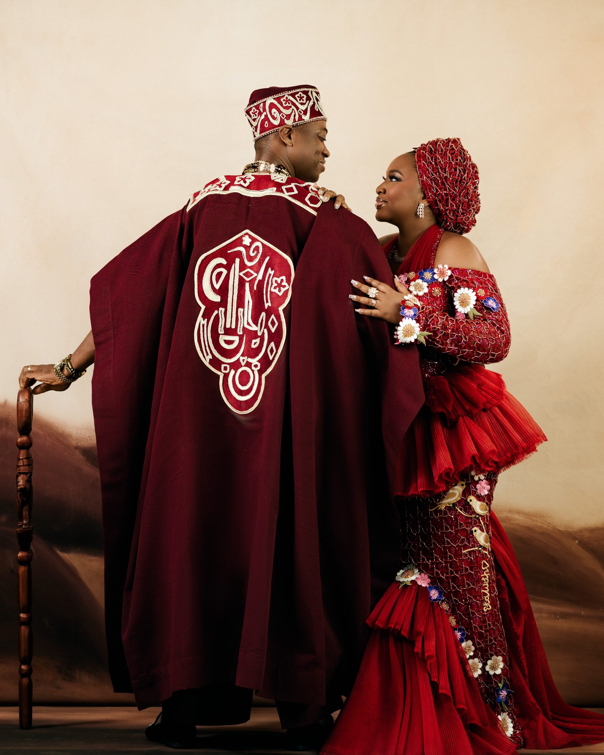 Back view of Lateef Adedimeji's embroidered burgundy Agbada and Mo Bimpe's off-the-shoulder red beaded gown with a pleated side drape.