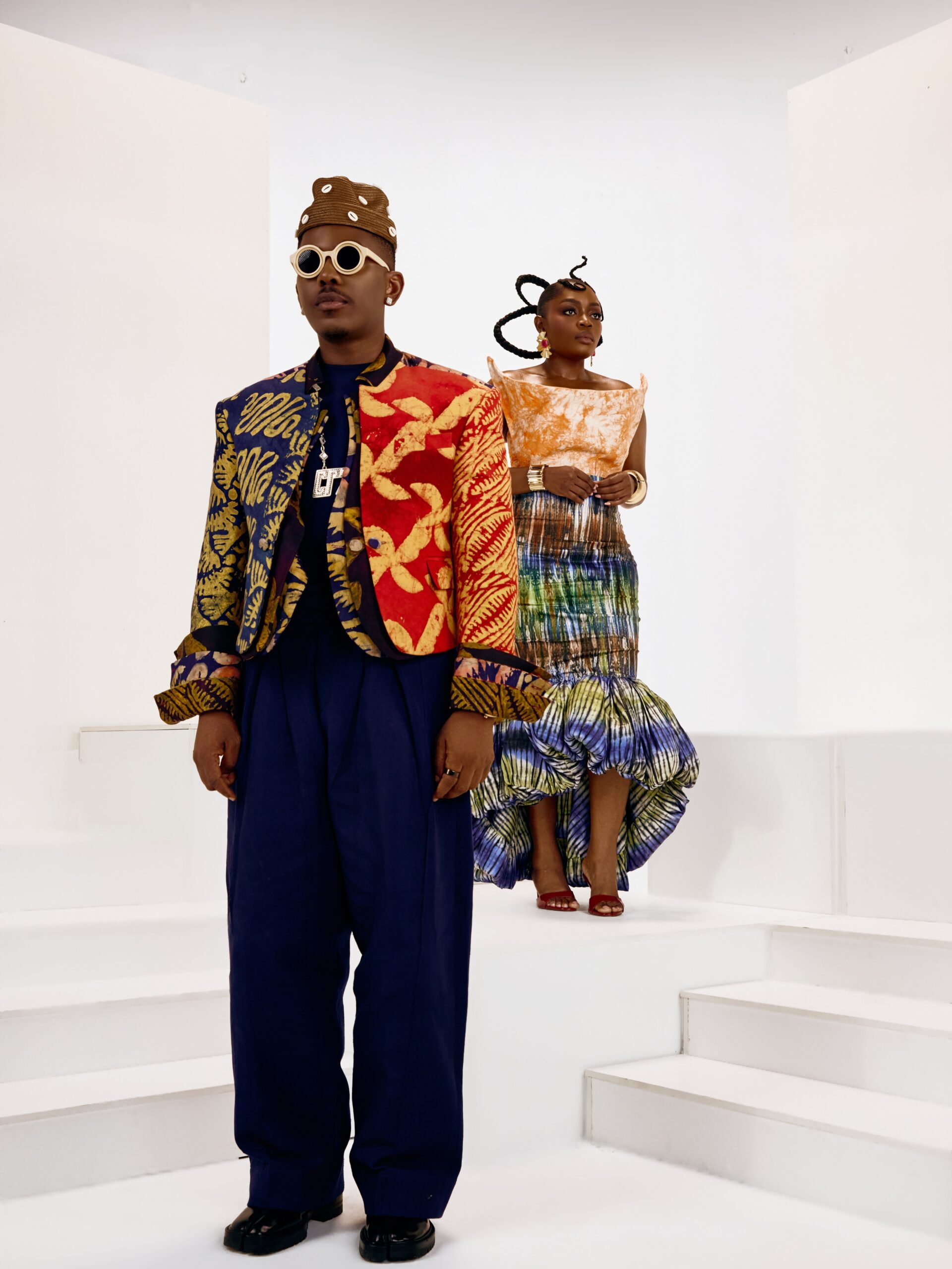 Timini Egbuson in a split-colour batik blazer standing on stairs in front of Bisola for The Four movie cast announcement.
