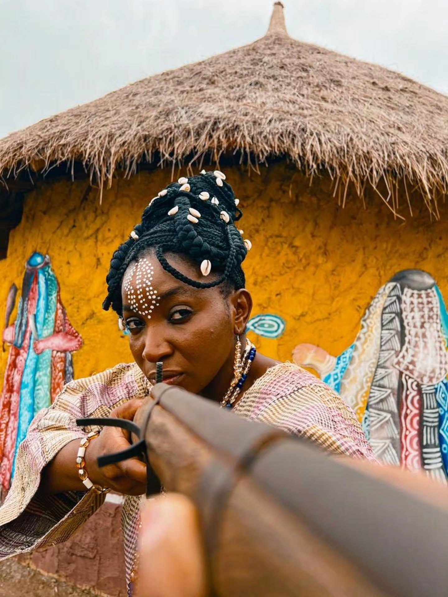 Omowunmi Dada aiming a rifle with cowrie-decorated braids and facial markings in Alahun The Weaver.