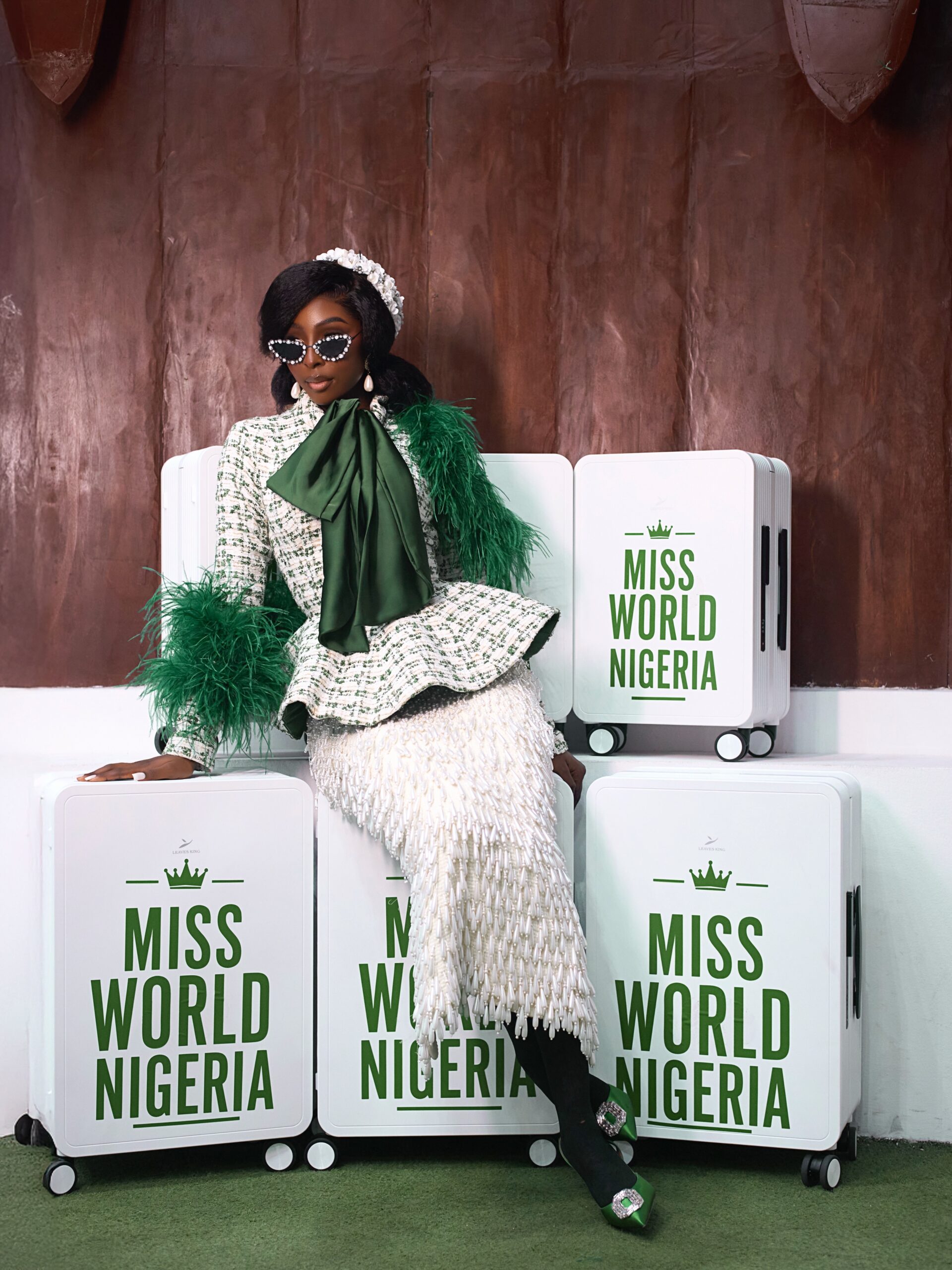 Tamunosoye Karibi-George wearing a green-and-white tweed outfit with green feather trim and sunglasses, sitting on branded Miss World Nigeria suitcases.
