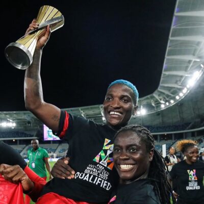 Tabitha Chawinga in locs carrying her sister Temwa Chawinga, who holds a trophy aloft wearing a FIFA Women's World Cup Qualified shirt.