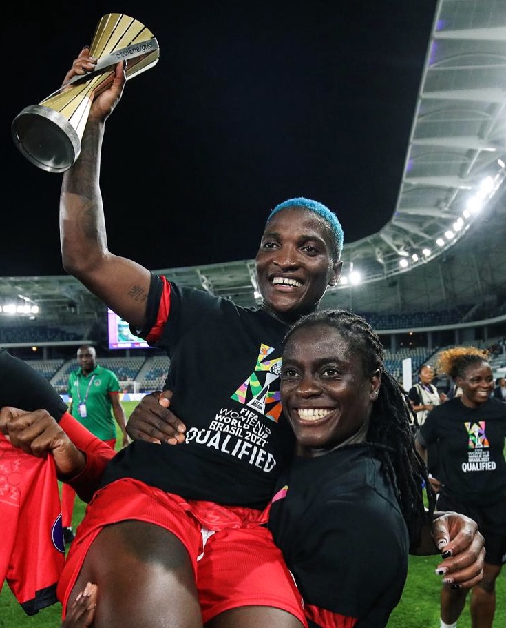 Tabitha Chawinga in locs carrying her sister Temwa Chawinga, who holds a trophy aloft wearing a FIFA Women's World Cup Qualified shirt.