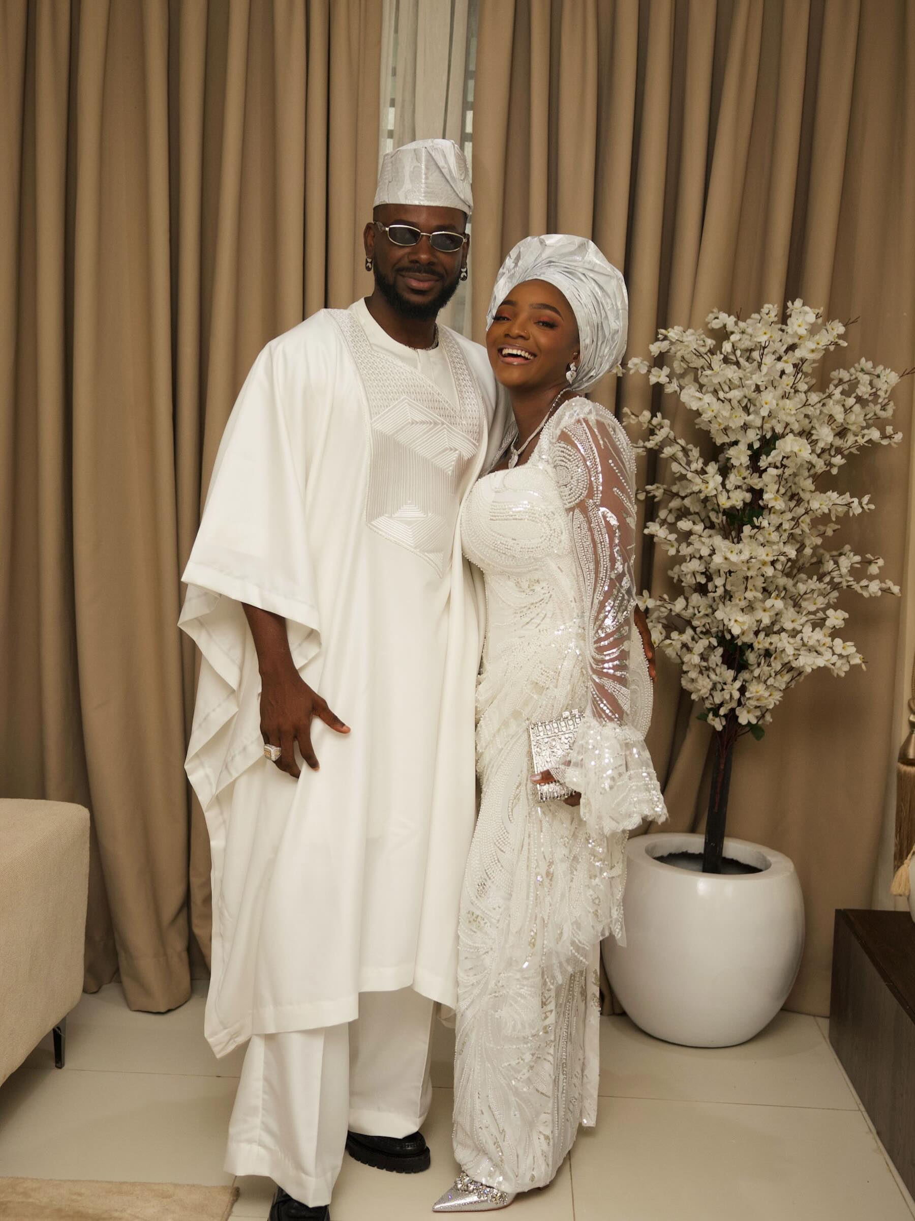 Simi and Adekunle Gold posing together in matching white traditional Yoruba outfits for their son Adeifemi's dedication.