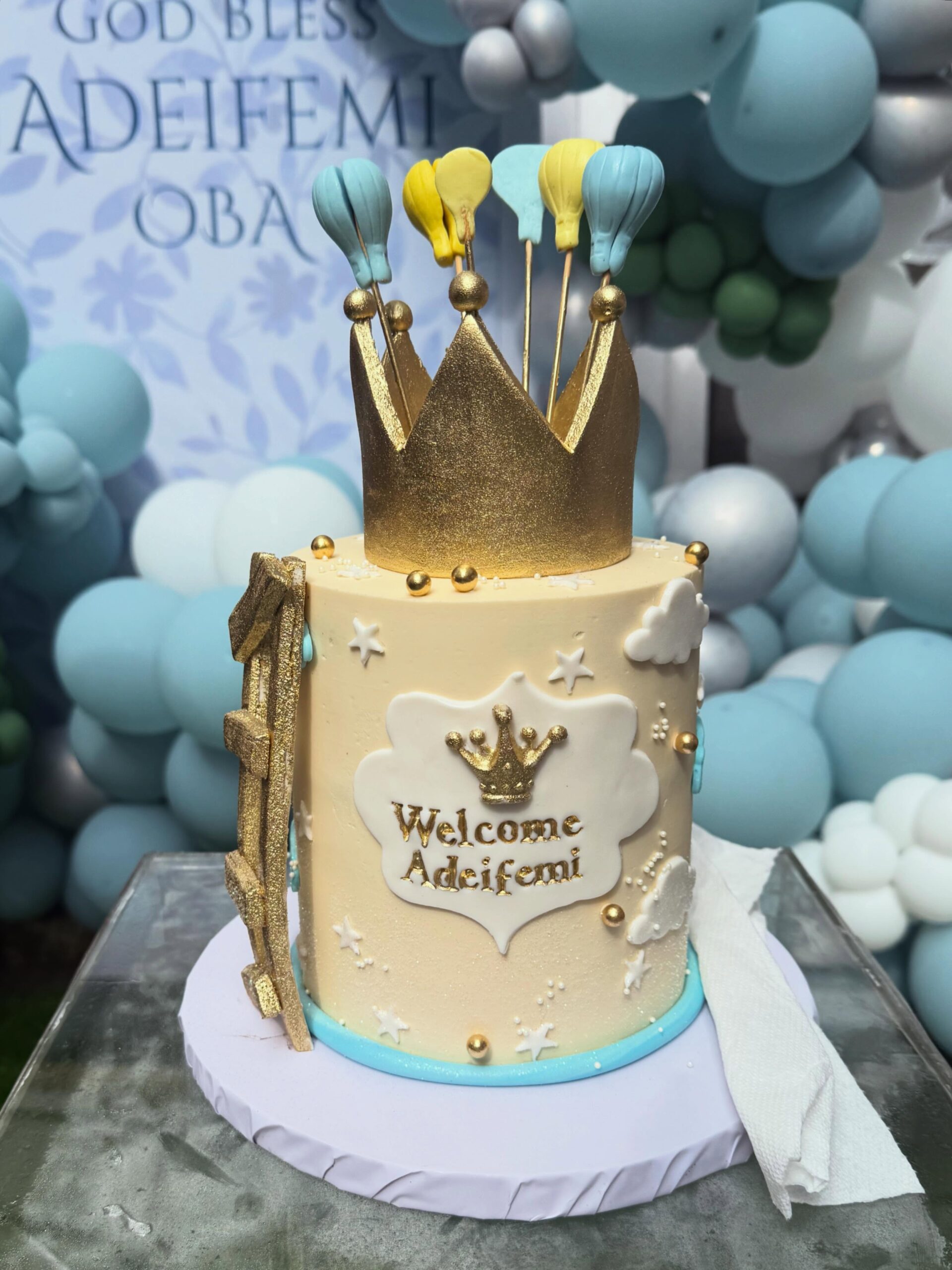 Custom celebration cake topped with a gold crown, air balloons, and "Welcome Adeifemi" inscription.