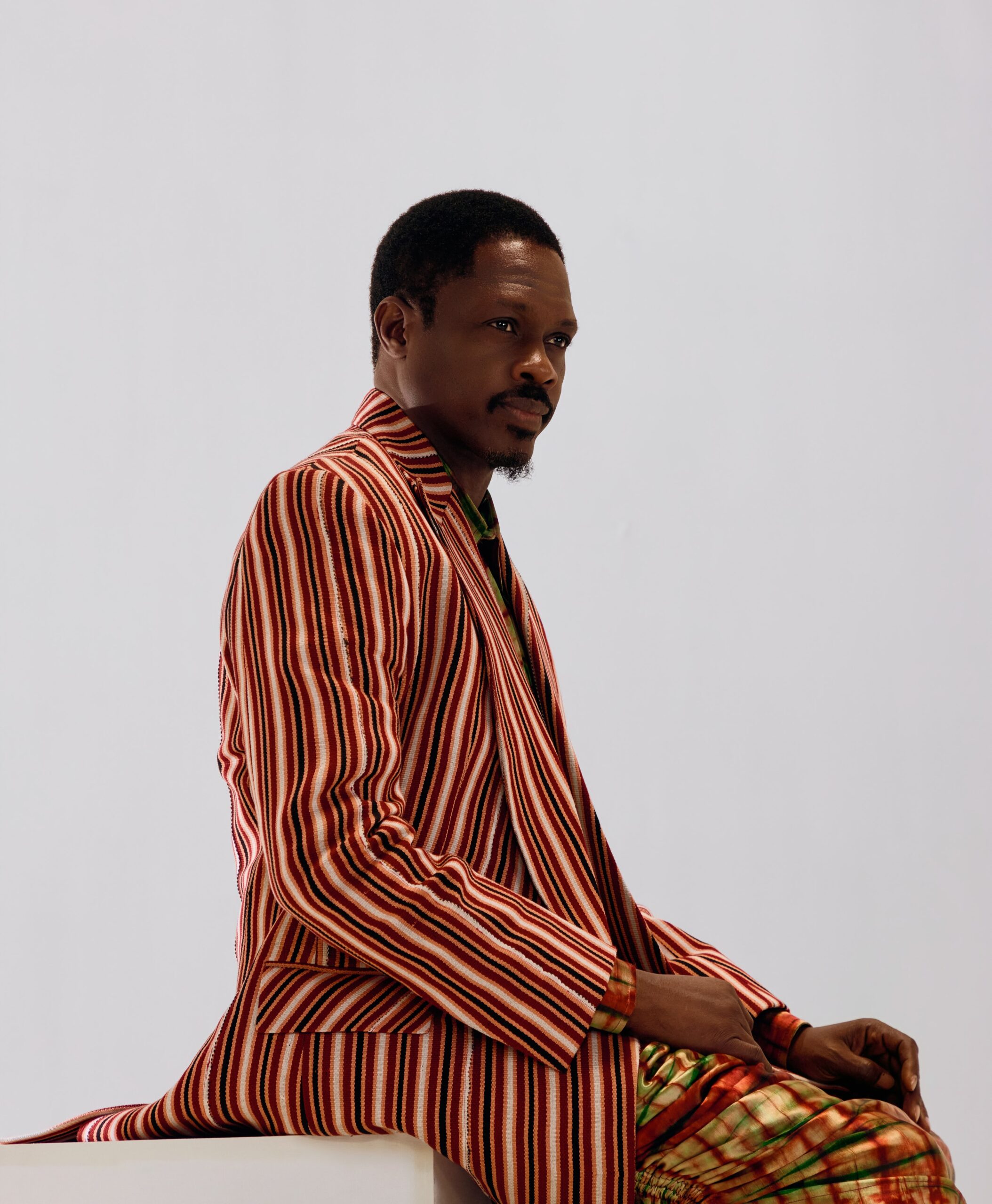 Actor Ali Nuhu posing in a red and cream striped longline jacket paired with green patterned trousers.