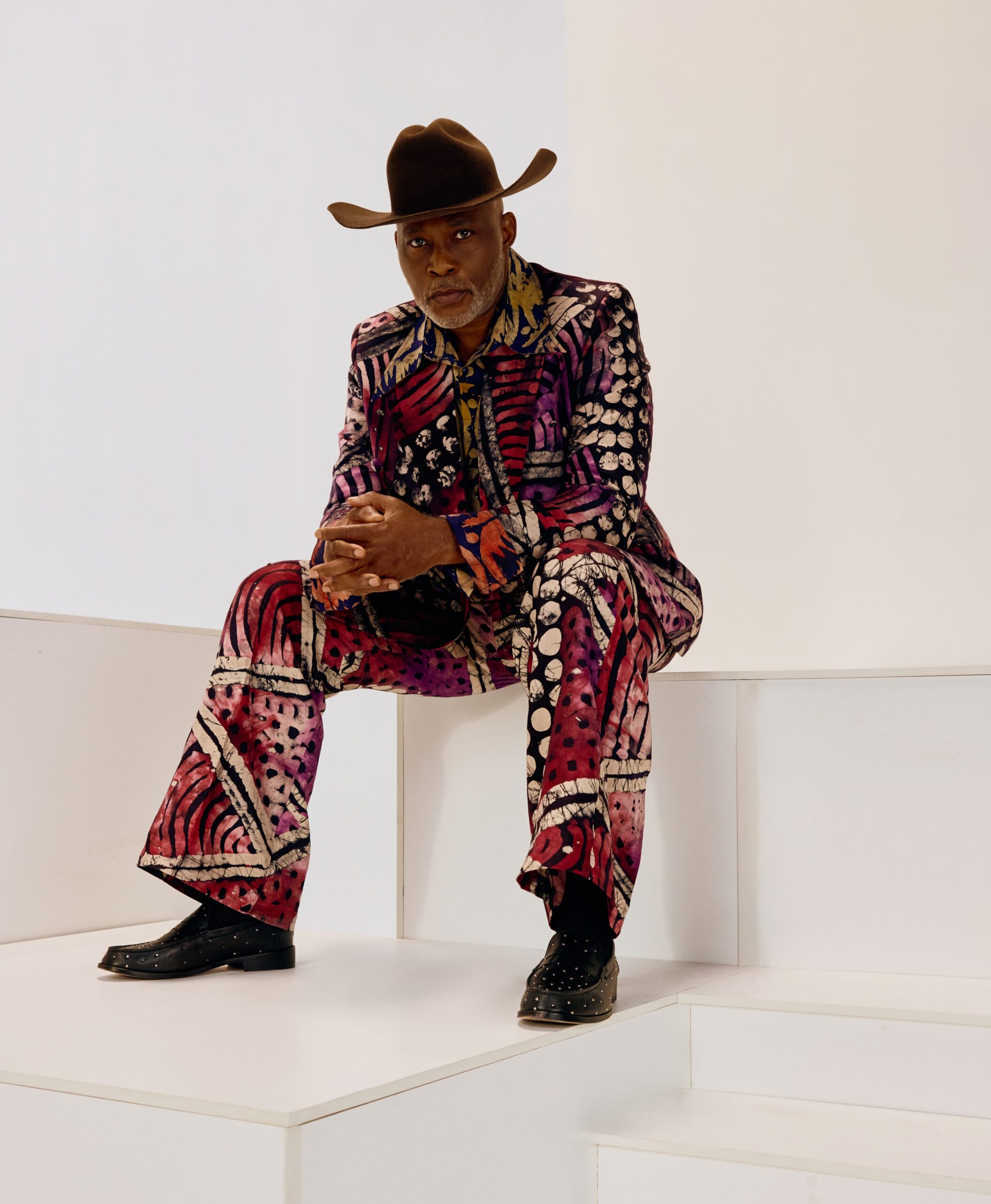 Actor Richard Mofe-Damijo seated on white blocks wearing a berry-patterned adire suit and brown cowboy hat for The Four movie launch.