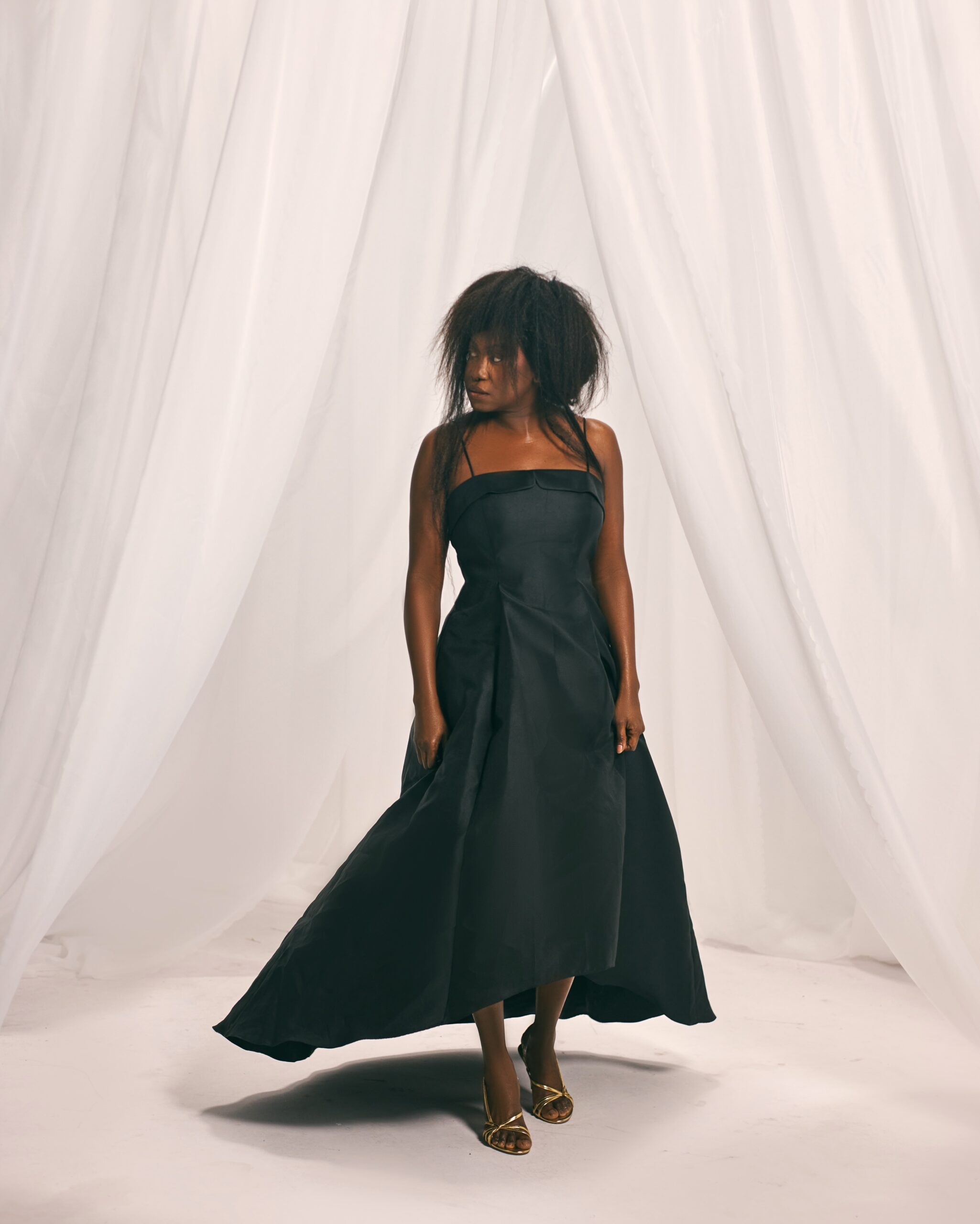 Niniola wearing a black flared dress set against draped white curtains in a still from her Raining O Dun Mi music video.