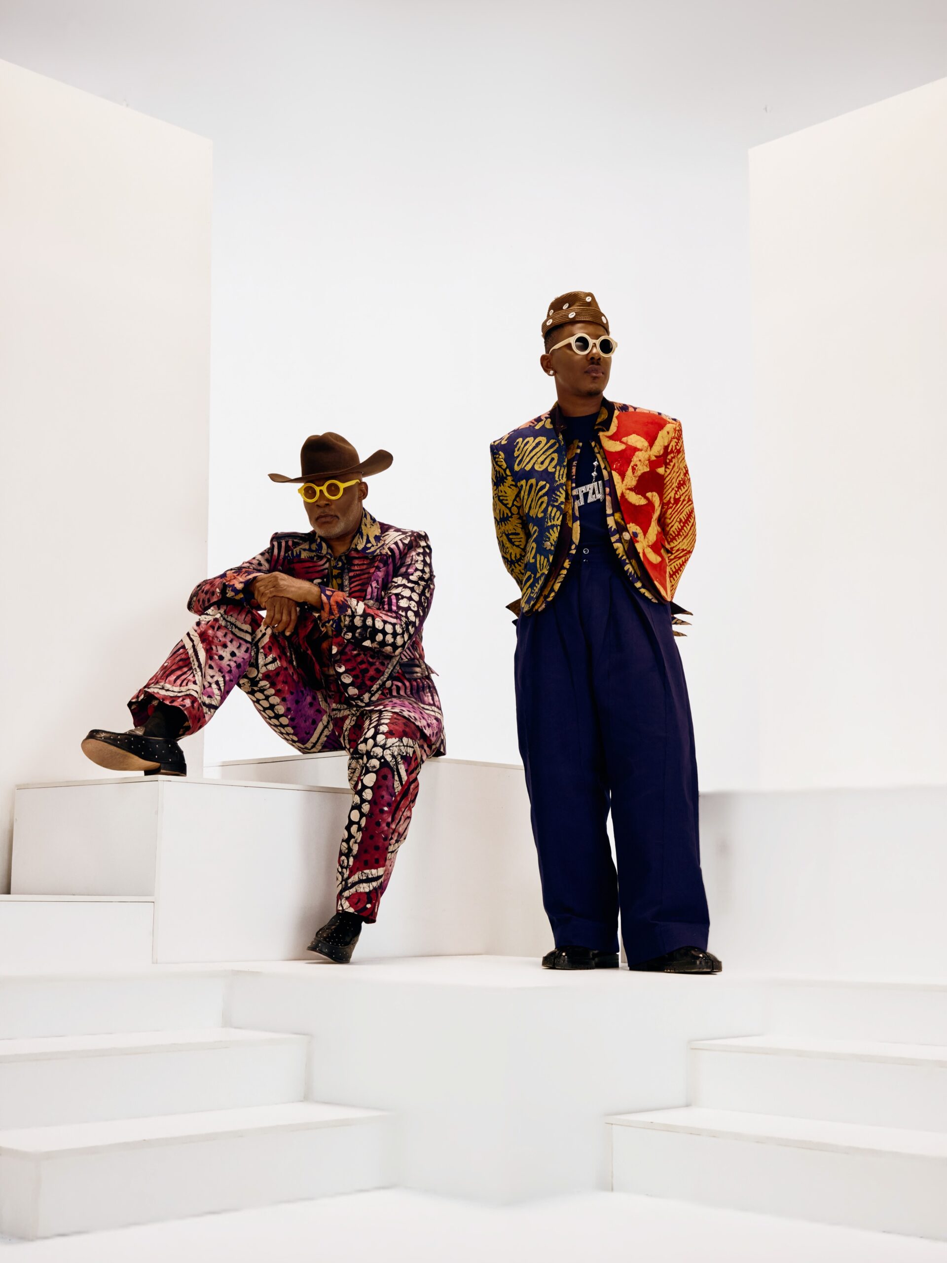 RMD seated in a purple adire suit alongside Timini Egbuson standing in a split-color blazer and wide-leg navy trousers on a white set.