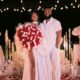 Meraiah Ekeinde in a white high-slit dress holding a red and white rose bouquet standing beside her fiancé Manny in all-white
