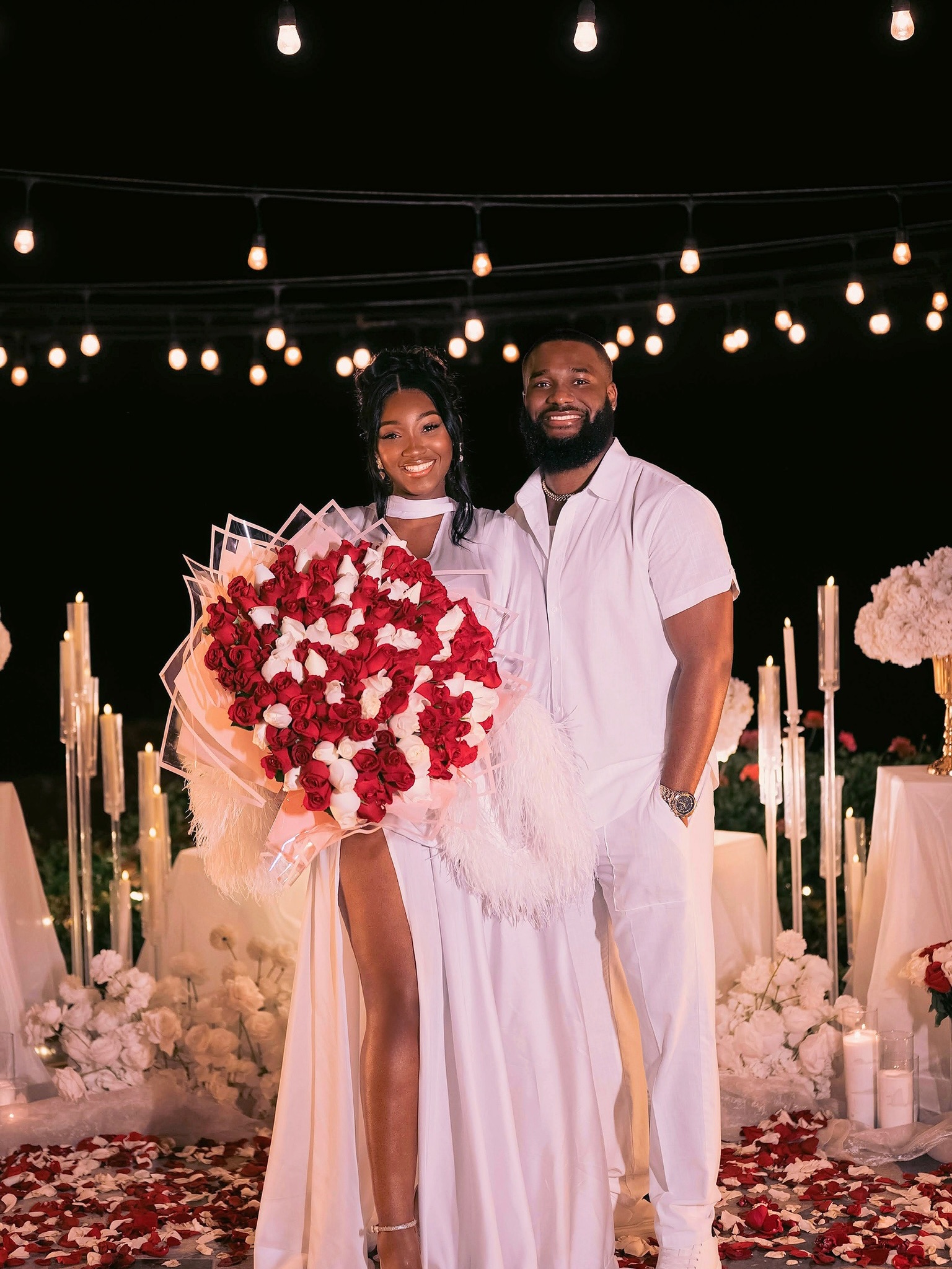 Meraiah Ekeinde in a white high-slit dress holding a red and white rose bouquet standing beside her fiancé Manny in all-white
