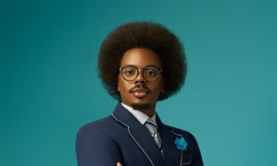 Enioluwa Adeoluwa wearing a navy blue suit with white contrast piping and eyeglasses against a turquoise background.