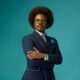 Enioluwa Adeoluwa wearing a navy blue suit with white contrast piping and eyeglasses against a turquoise background.