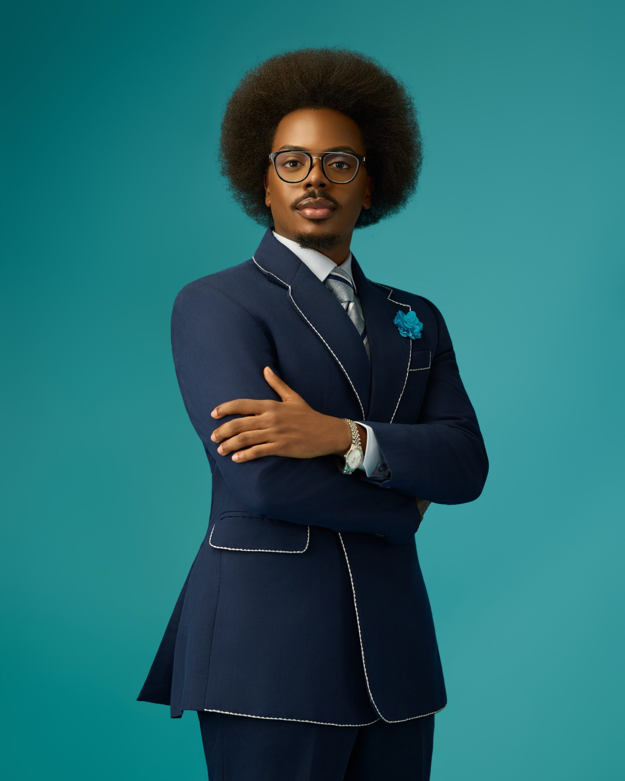 Enioluwa Adeoluwa wearing a navy blue suit with white contrast piping and eyeglasses against a turquoise background.