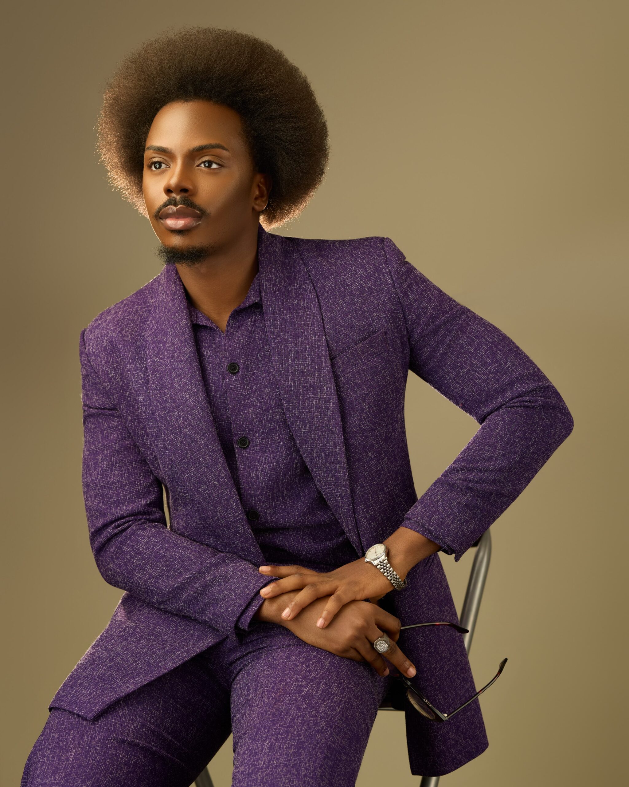 Enioluwa Adeoluwa sitting in a textured royal purple suit holding sunglasses with a rounded Afro hairstyle.