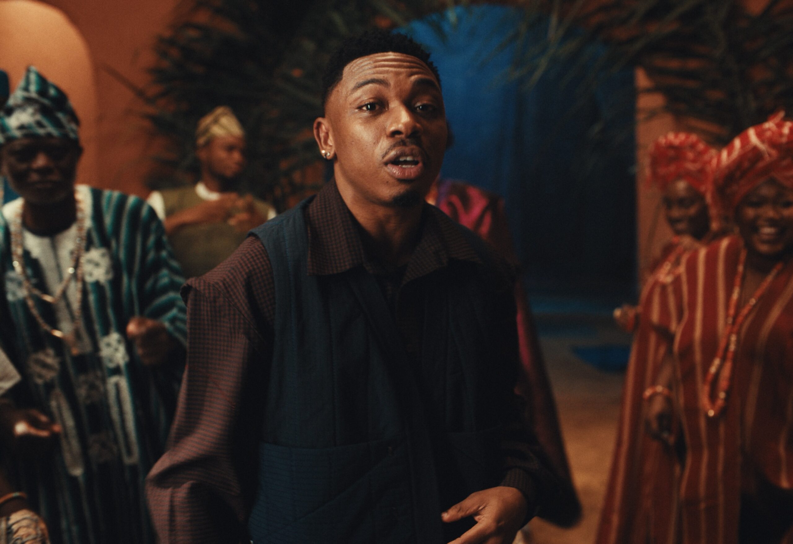 Mayorkun singing in a patterned vest and collared shirt with traditional drummers in the background.