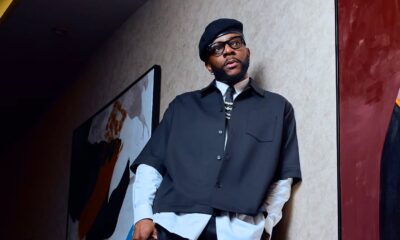 Ebuka Obi-Uchendu leaning against a wall in wide-leg cropped trousers, white crew socks, black loafers, and holding a Louis Vuitton monogram travel bag.