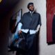 Ebuka Obi-Uchendu leaning against a wall in wide-leg cropped trousers, white crew socks, black loafers, and holding a Louis Vuitton monogram travel bag.