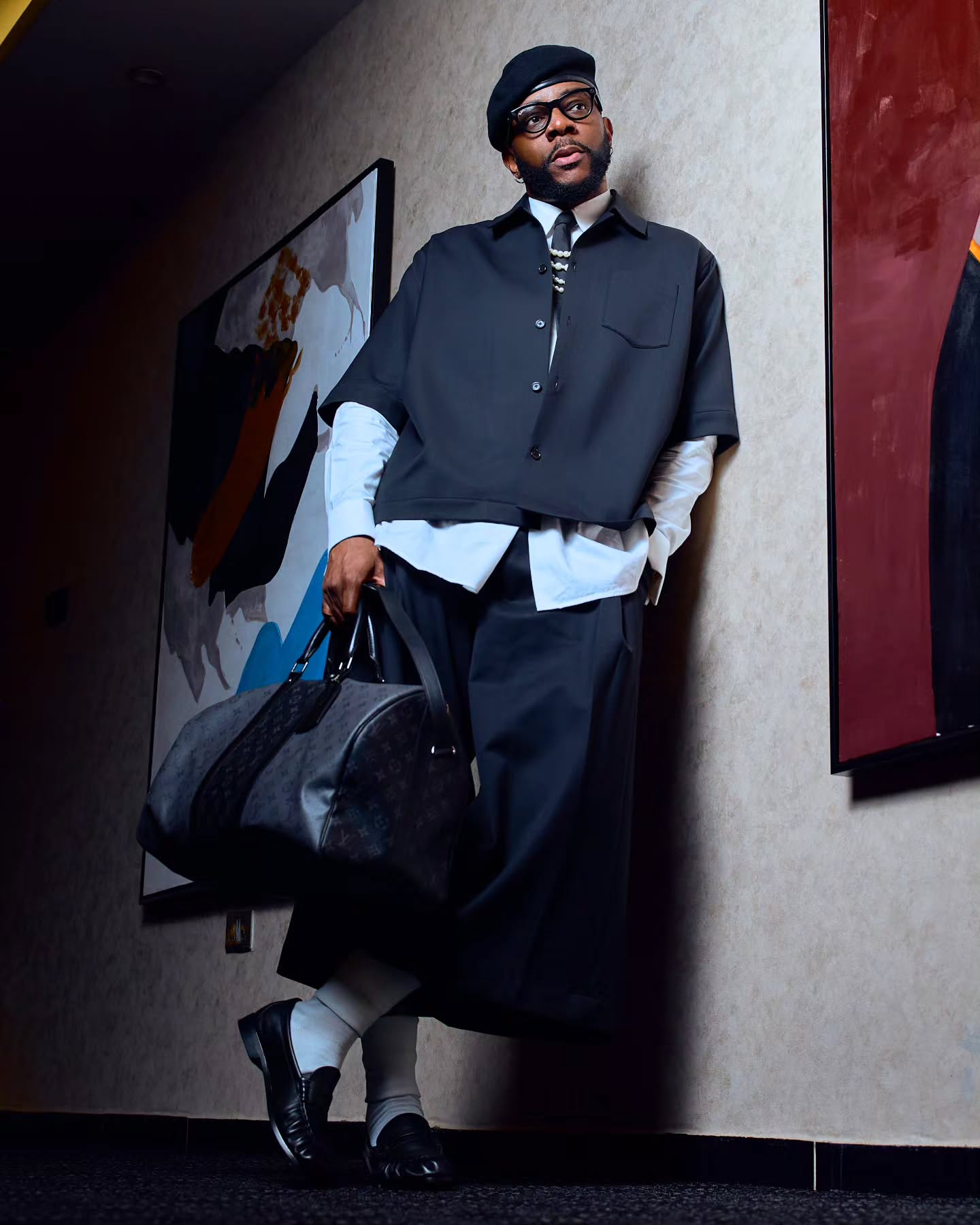 Ebuka Obi-Uchendu leaning against a wall in wide-leg cropped trousers, white crew socks, black loafers, and holding a Louis Vuitton monogram travel bag.