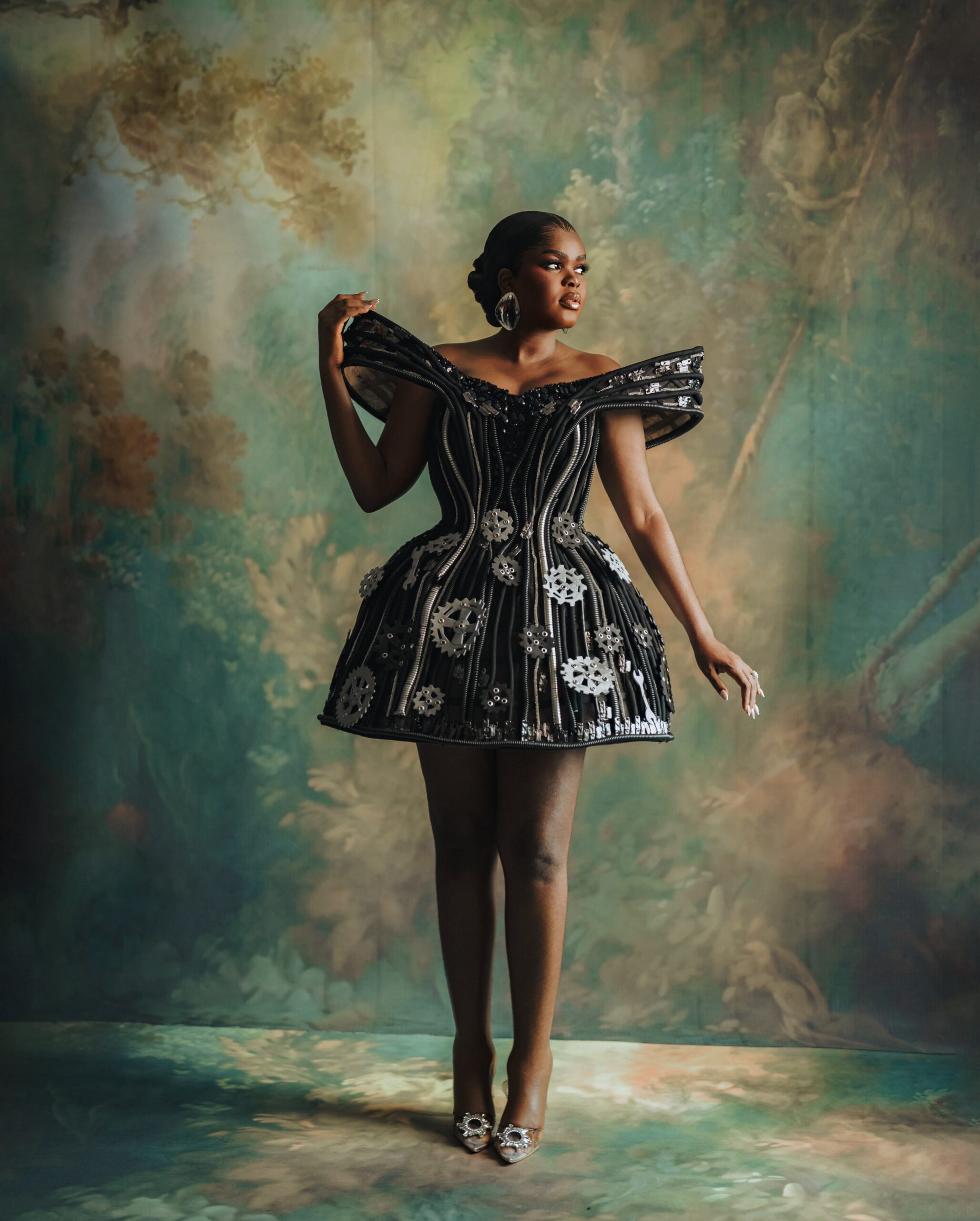 Jarvis posing solo in a black A-line mini dress adorned with gears, cogs, spanners, and exposed zips.
