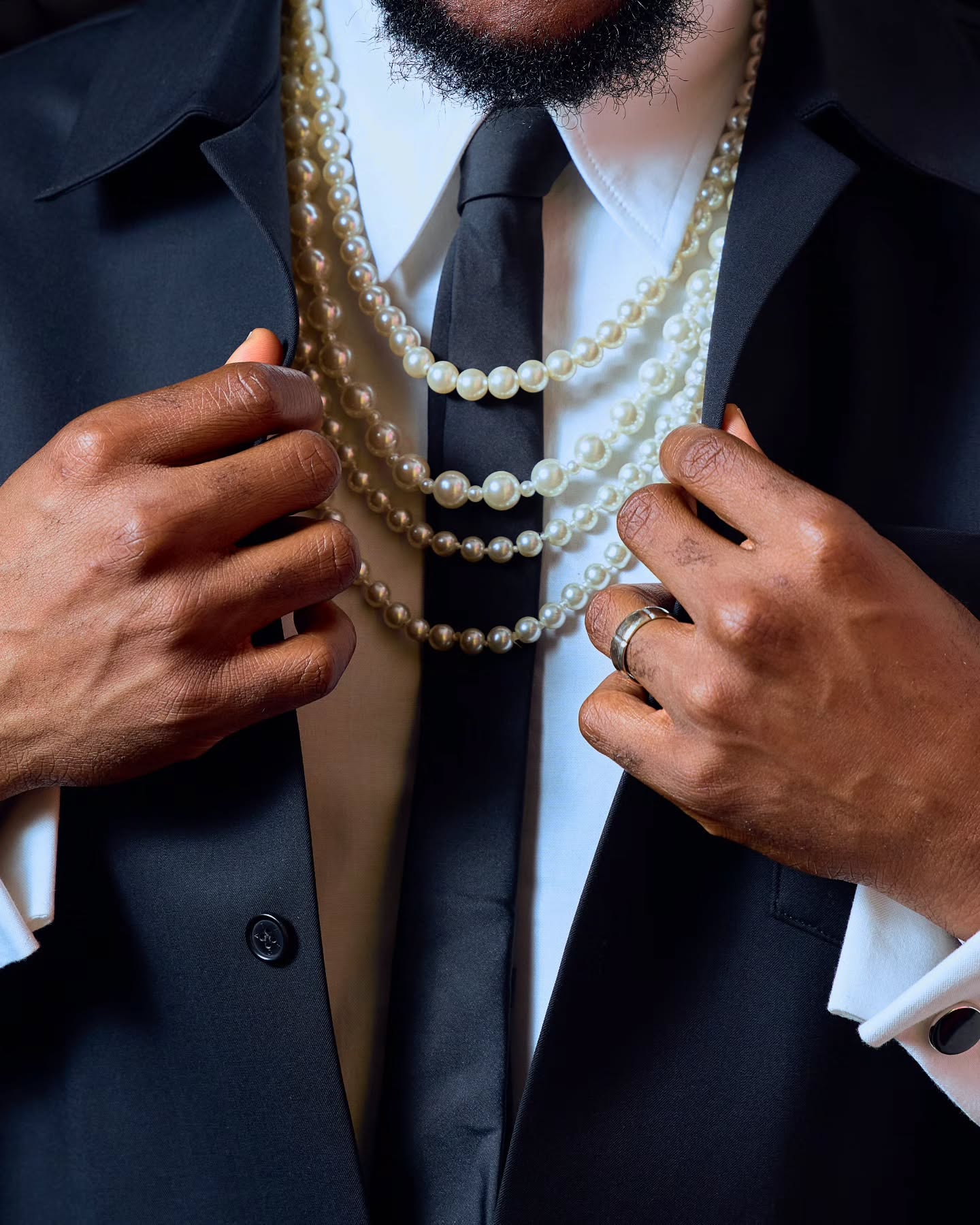 Extreme close-up of Ebuka Obi-Uchendu adjusting a black button-down shirt to reveal a multi-strand pearl necklace draped over a black tie.