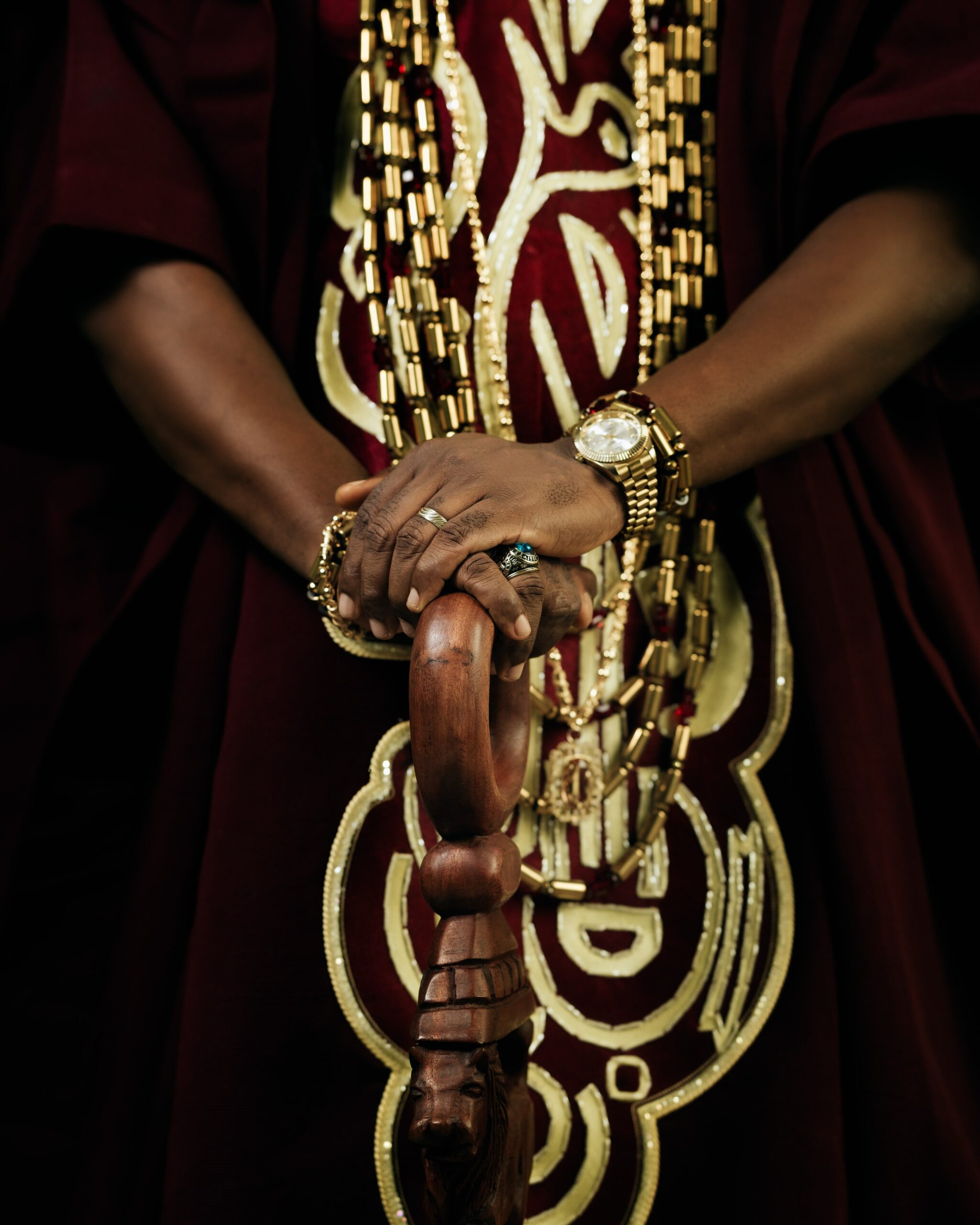 Close-up of Adedimeji Lateef's hands resting on a carved wooden staff, showing layered gold beads, rings, and a gold wristwatch.