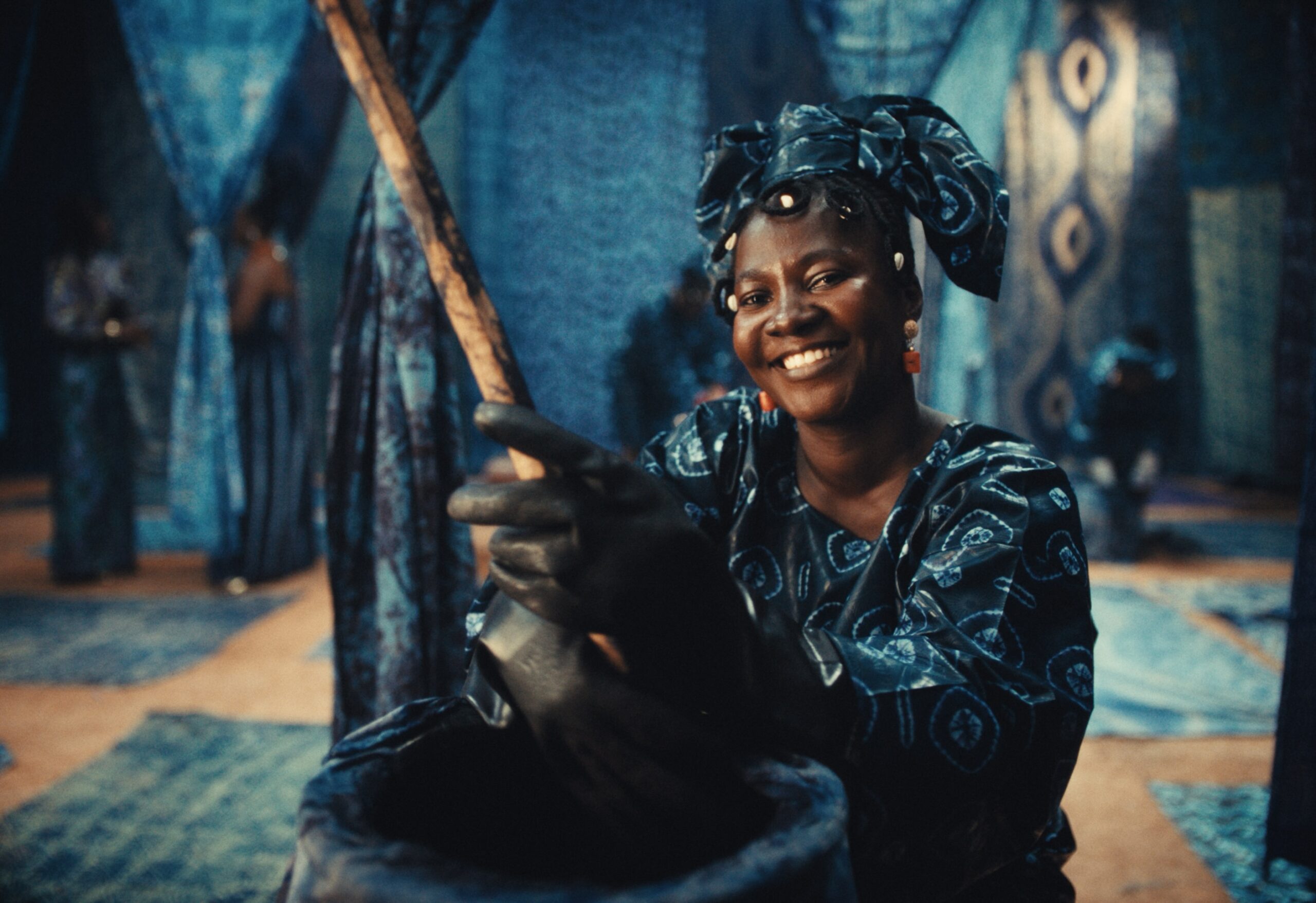 Smiling woman in indigo Adire headwrap and outfit holding a wooden pestle in an Adire workshop.