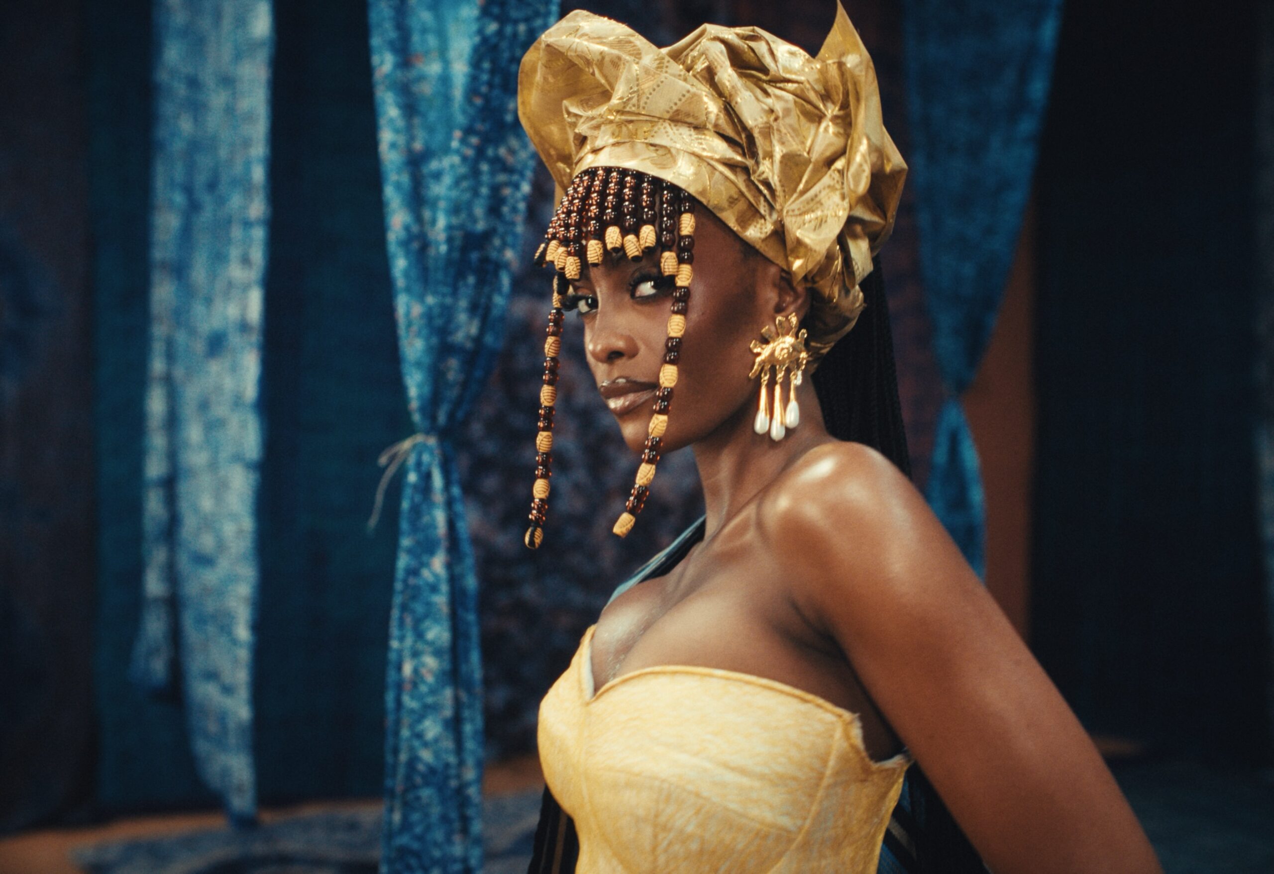 Model wearing a structured gold headwrap with beaded fringe and statement gold earrings against blue Adire drapes.