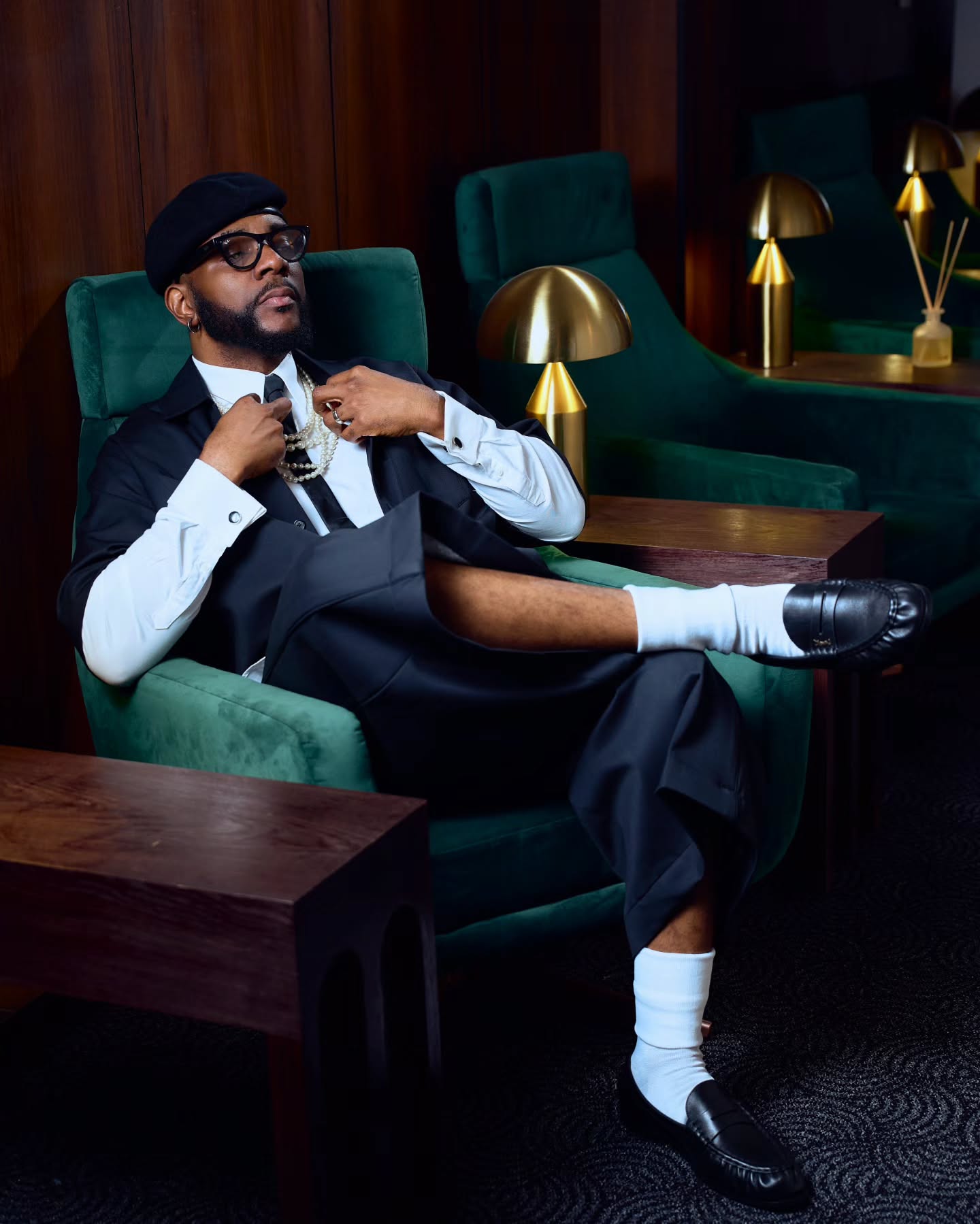 Ebuka Obi-Uchendu seated in a green velvet armchair with crossed legs, showcasing white crew socks, black loafers, and a beret.