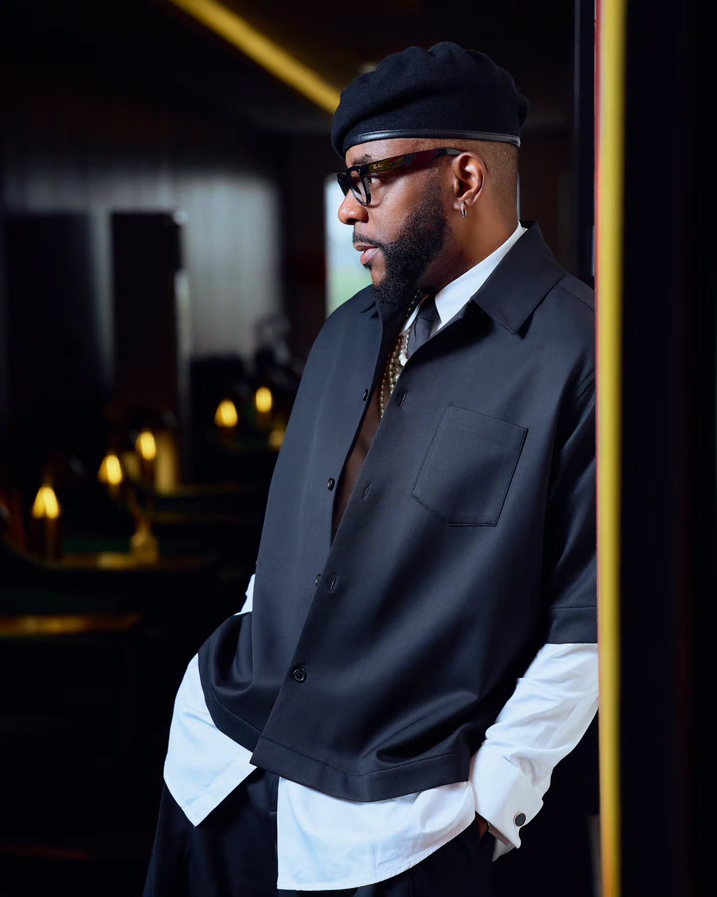 Close-up profile of Ebuka Obi-Uchendu wearing a black beret, black-framed glasses, and a layered collar with a tie and pearl necklace.