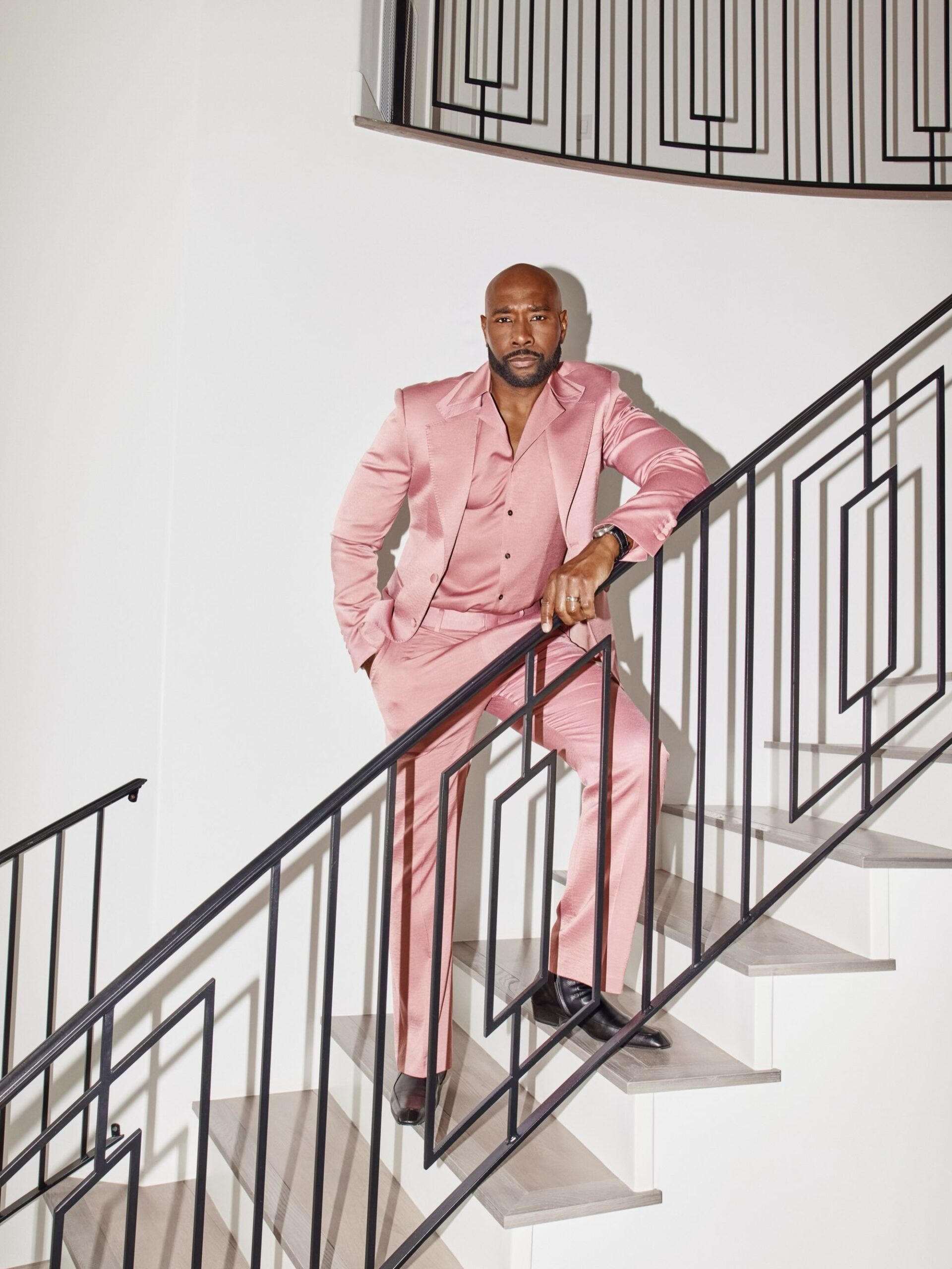 Morris Chestnut standing on a modern staircase in high-waisted rose pink trousers and black leather dress shoes.
