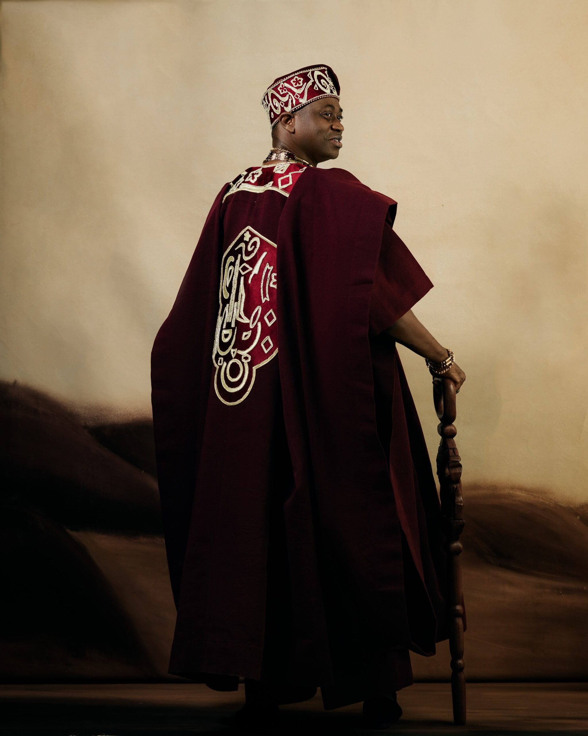 Rear view of Adedimeji Lateef in a deep wine Agbada by Tochini showing intricate gold back embroidery and holding a carved walking stick.