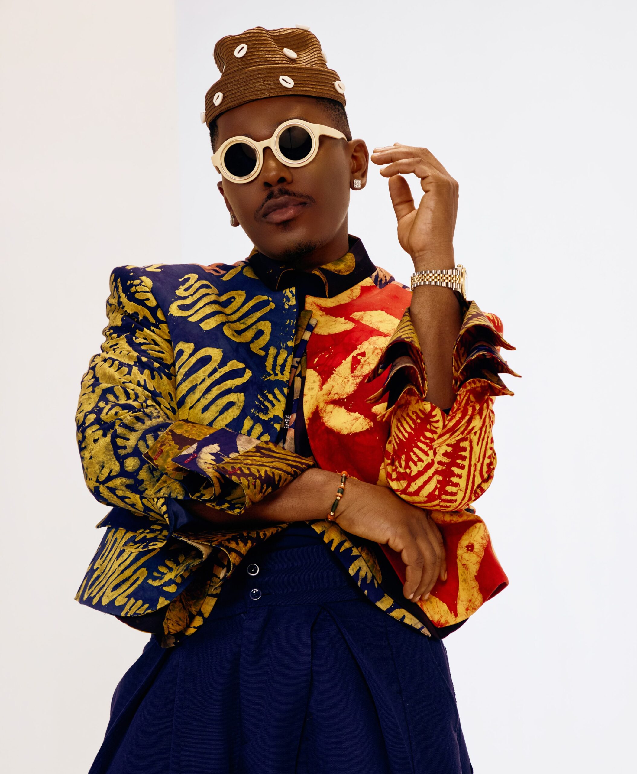 Timini Egbuson wearing a multi-coloured patterned jacket, dark trousers, cowrie-shell hat, and round sunglasses.
