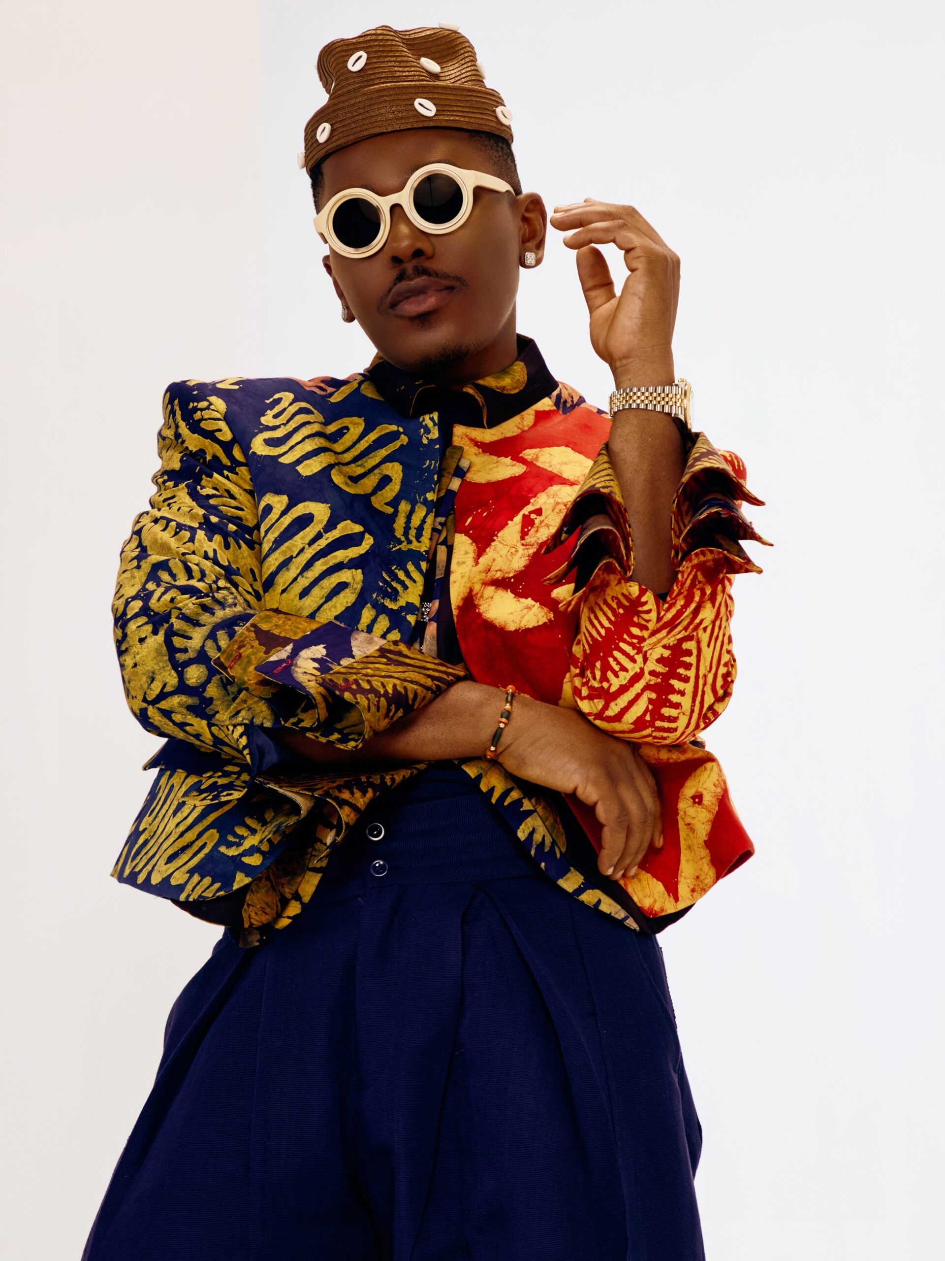 Timini Egbuson posing in a custom split-color adire jacket, cowrie shell cap, and off-white sunglasses.