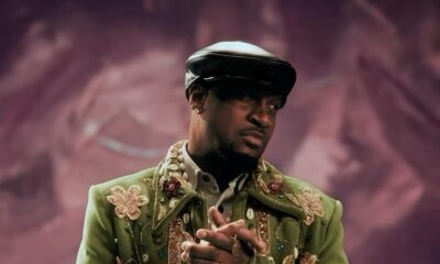 Mr P Peter Okoye wearing a green floral-embroidered jacket, cowrie shell embellishments, and a black cap with hands clasped.