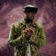 Mr P Peter Okoye wearing a green floral-embroidered jacket, cowrie shell embellishments, and a black cap with hands clasped.