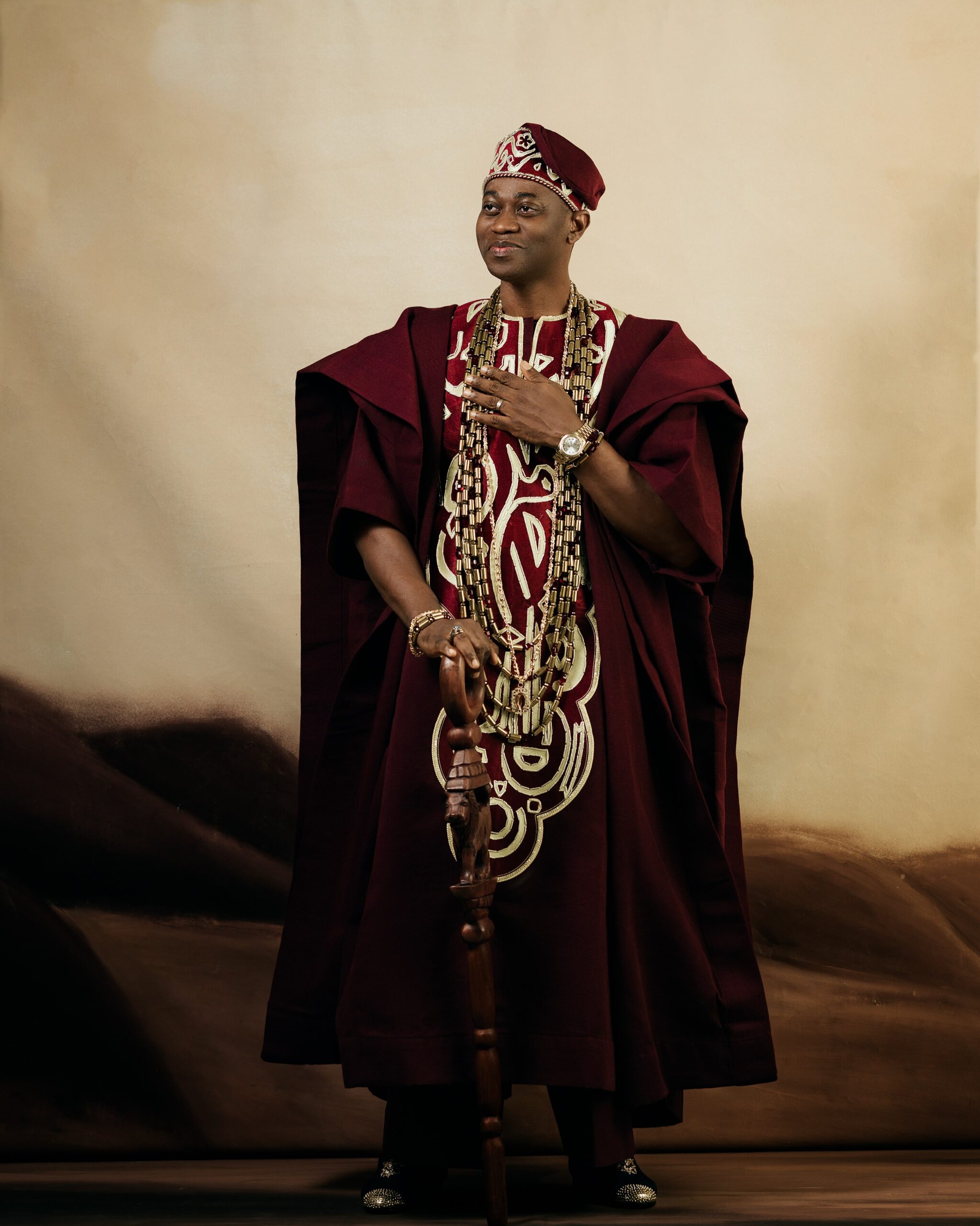 Adedimeji Lateef with his hand on his chest wearing a gold-embroidered wine Agbada, matching fila, and traditional coral beads.