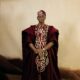 Full-length studio portrait of Adedimeji Lateef in a wine three-piece Agbada set, black velvet loafers, and coral jewellery.